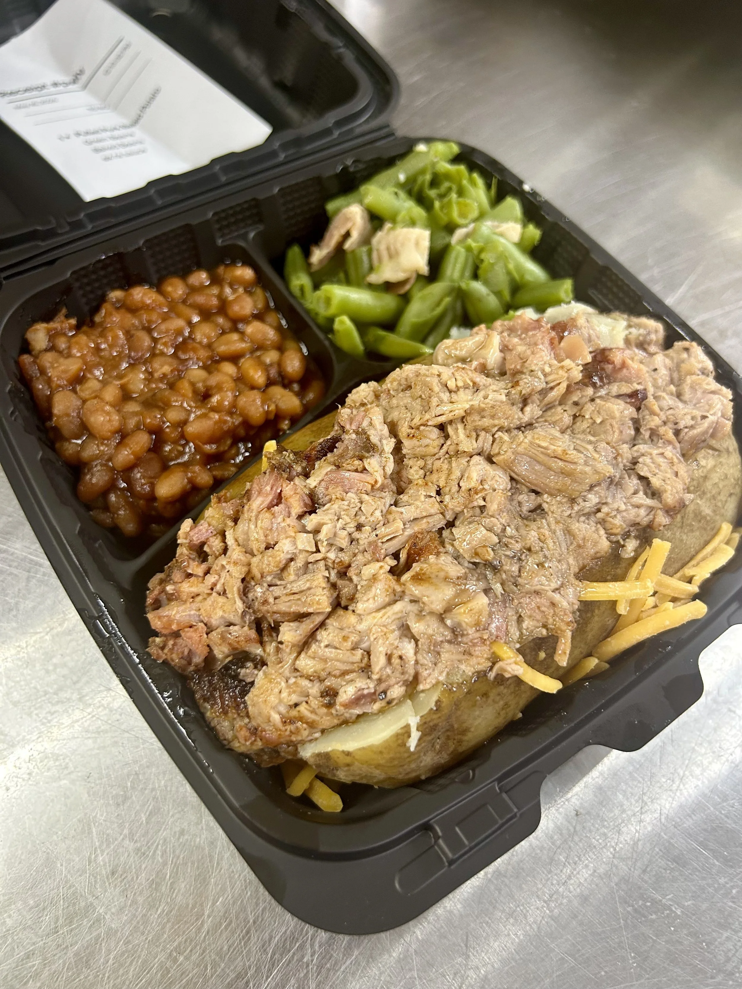 A black meal prep container with a baked potato loaded with chopped brisket, green beans, baked beans, and shredded cheese, placed on a stainless steel surface.