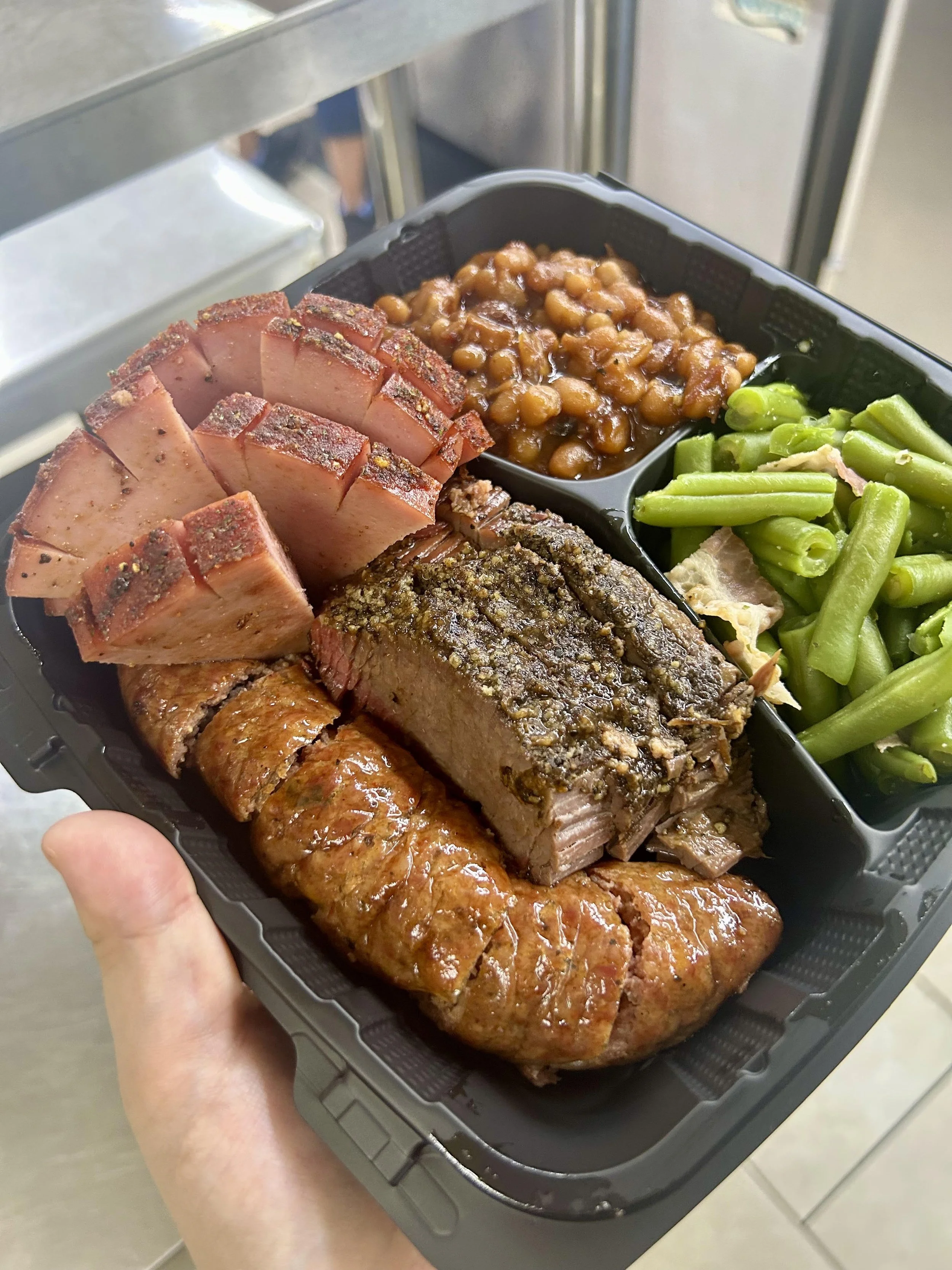 A hand holding a black tray filled with sliced cooked meats and baked beans, in the background a gray metallic kitchen surface.
