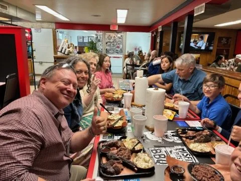Family enjoying a meal at a restaurant table with plates of barbecue, sides, and drinks, smiling and giving thumbs up.