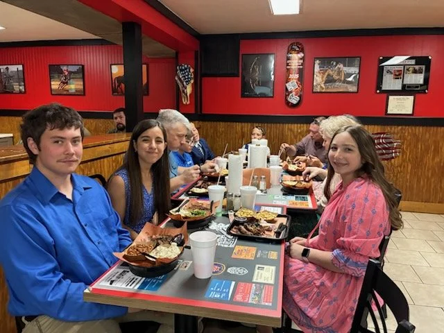 A group of young people sitting around a table in a casual restaurant, enjoying a meal with trays of food and drinks, decorated with American flags on the wall.
