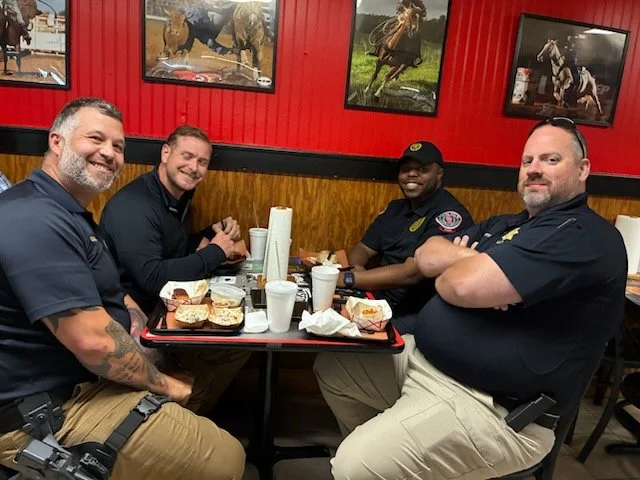 Four men sitting at a restaurant table with food, drinks, and paper towels, smiling at the camera. The background features red walls and photographs of horses.