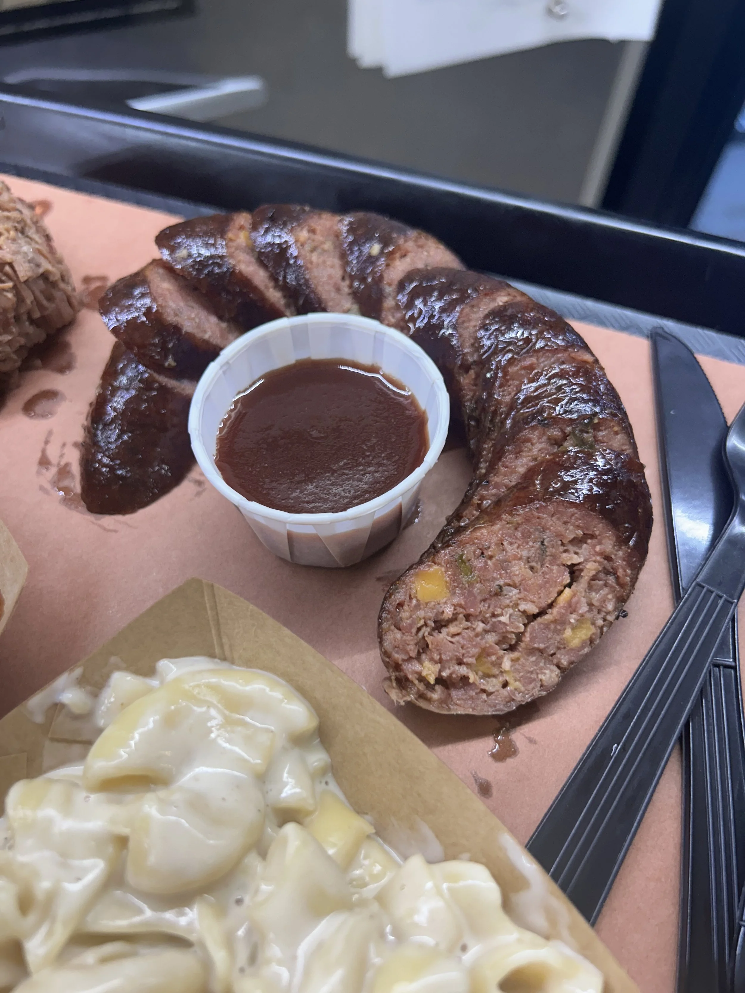 Barbecue sausage link sliced open with a cup of barbecue sauce on a tray with a serving of potato salad.