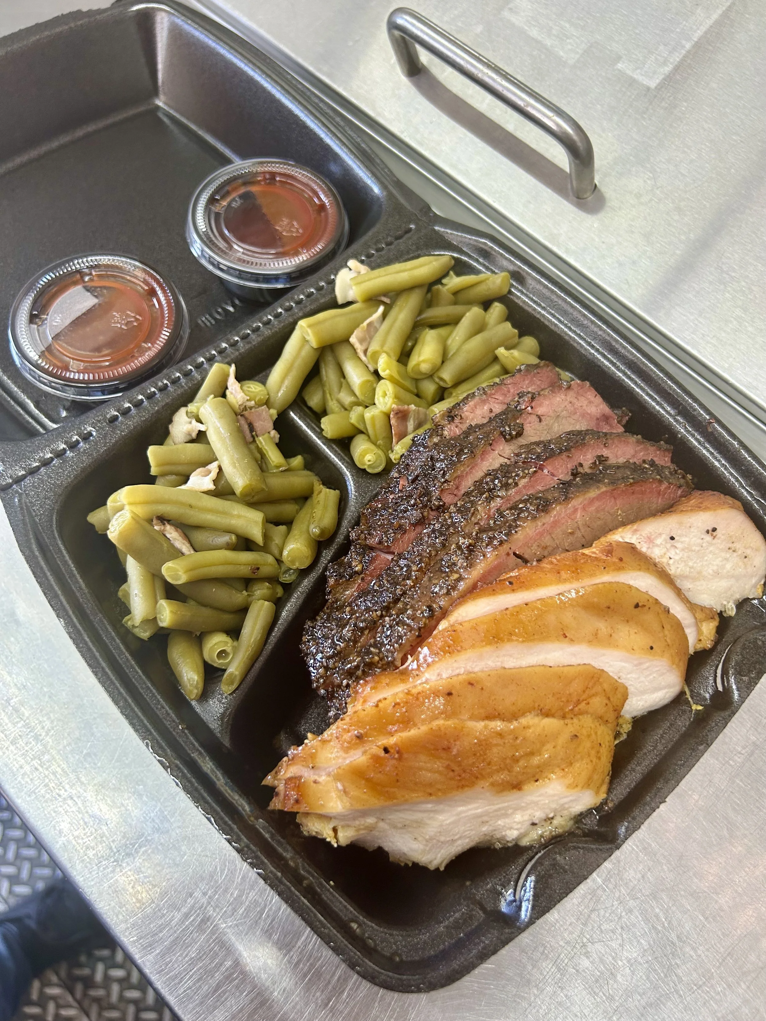 A black to-go meal tray holding sliced brisket, sliecd smoked chicken breast, garlic bacon green beans, and two containers of sauce.