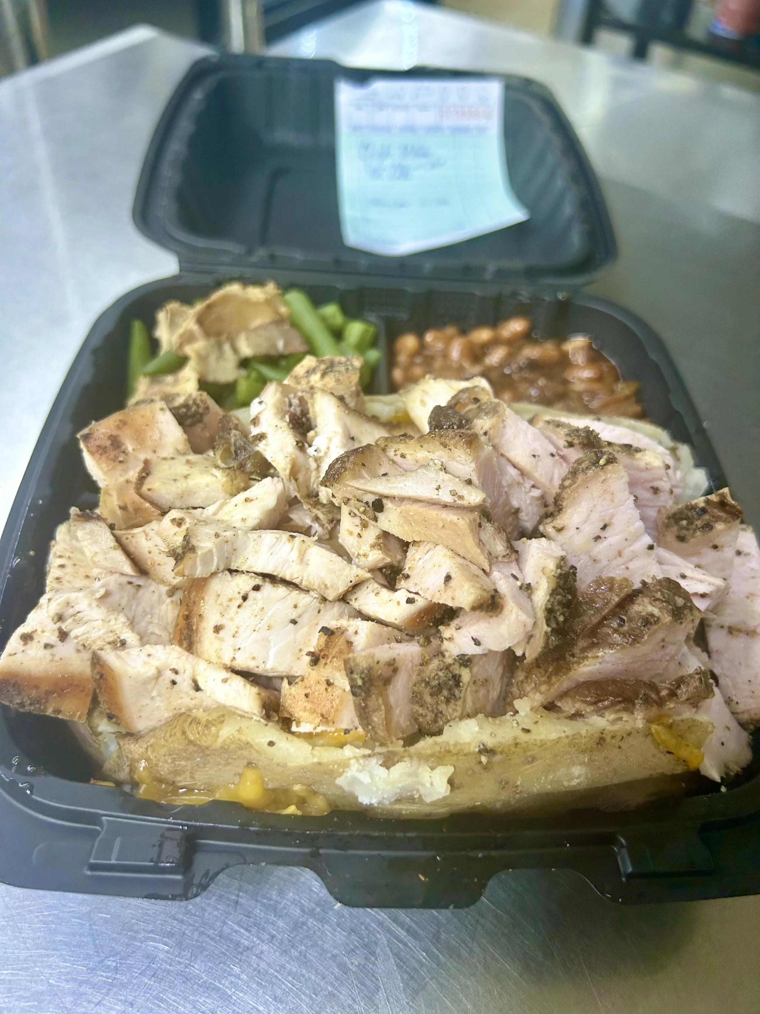 Close-up of a meal with sliced smoked chicken breast, served with green beans and baked beans in a black plastic takeout container.
