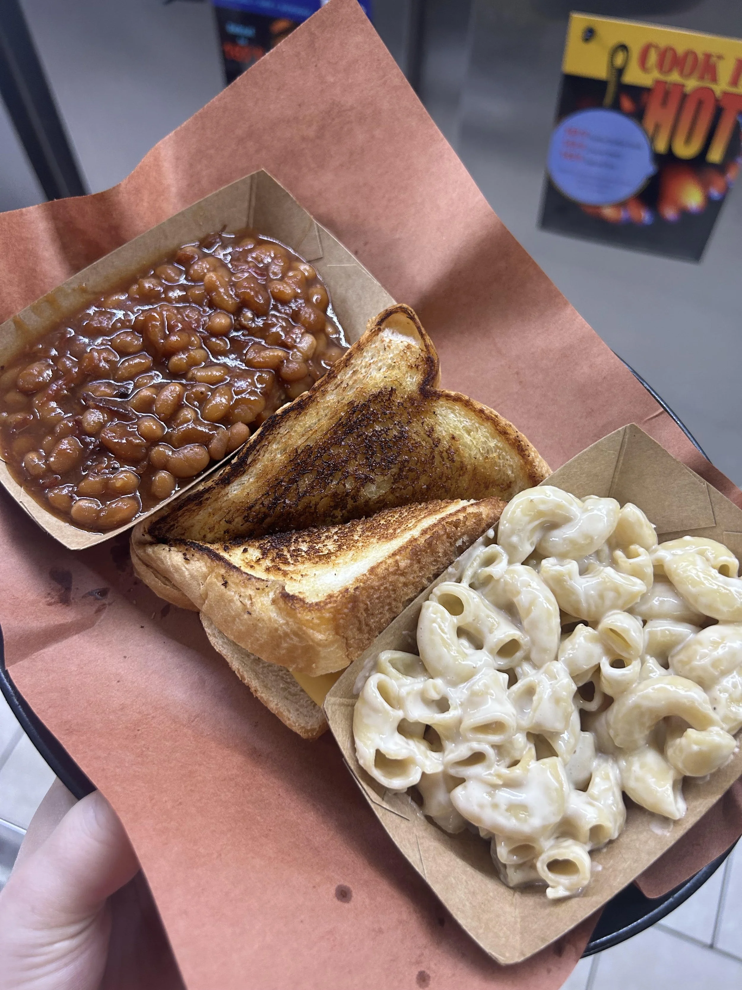 Hand holding a tray with baked macaroni and cheese, toasted sandwich, baked beans in a paper tray, and potato salad, in a casual eatery setting.