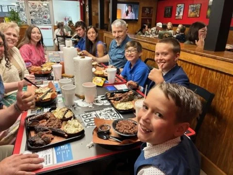 Family and friends gathered around a table in a restaurant enjoying a meal with barbecue dishes, drinks, and desserts.