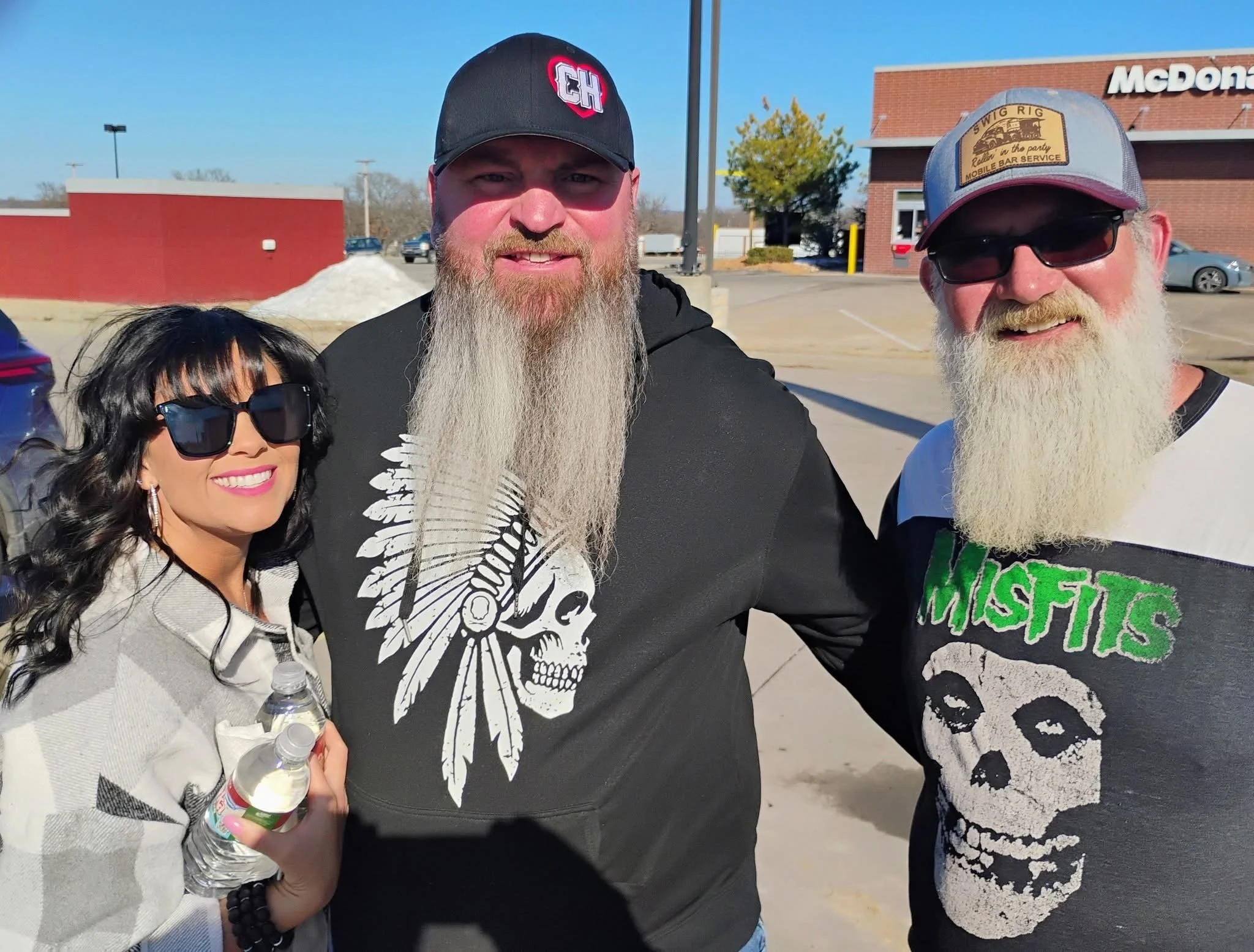 A group of four people outdoors near a McDonald's restaurant, smiling at the camera. Two men have long white beards and are wearing caps, one with a black cap featuring a red and white logo and another with a gray cap that has a yellow patch. The wom