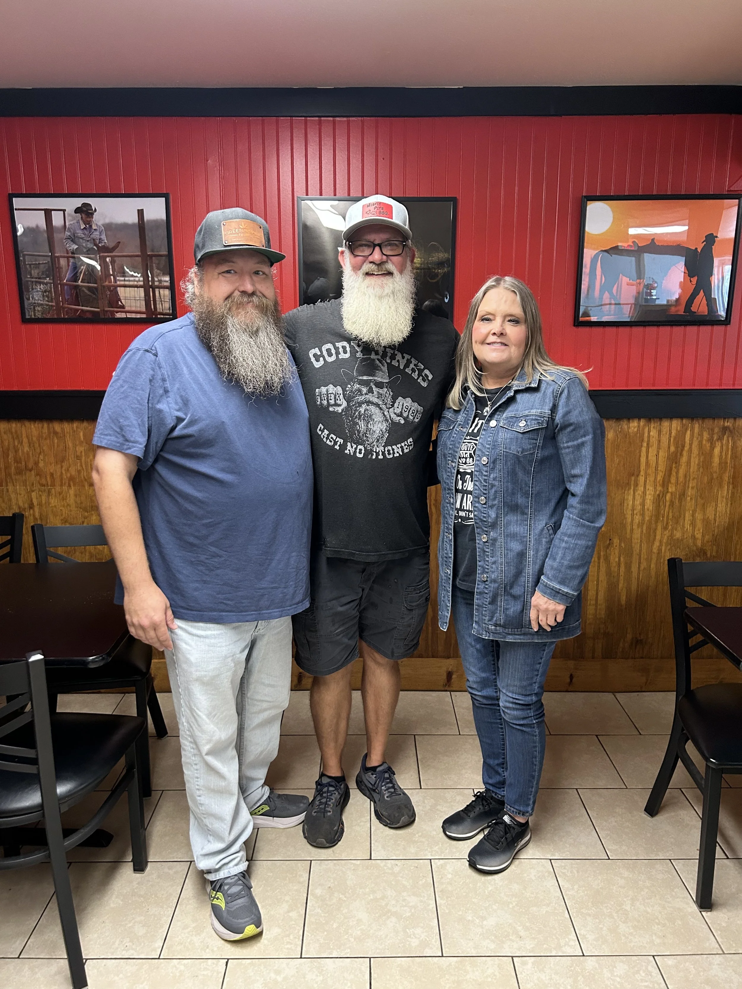 Three people standing together inside a restaurant with red walls and framed pictures, smiling and posing for the photo.