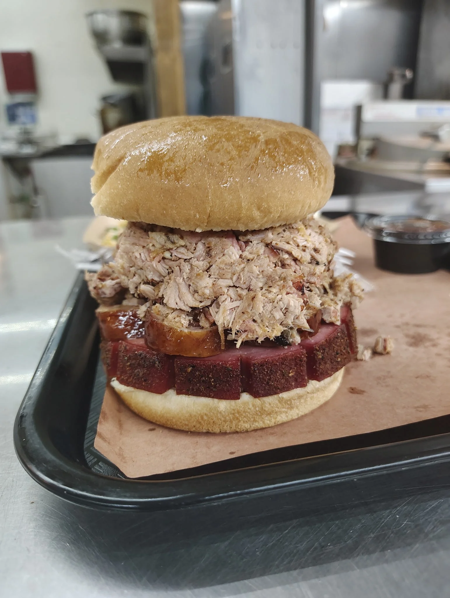 Close-up of an oversized sandwich with pulled pork and bologna on a bun, placed on a black tray.