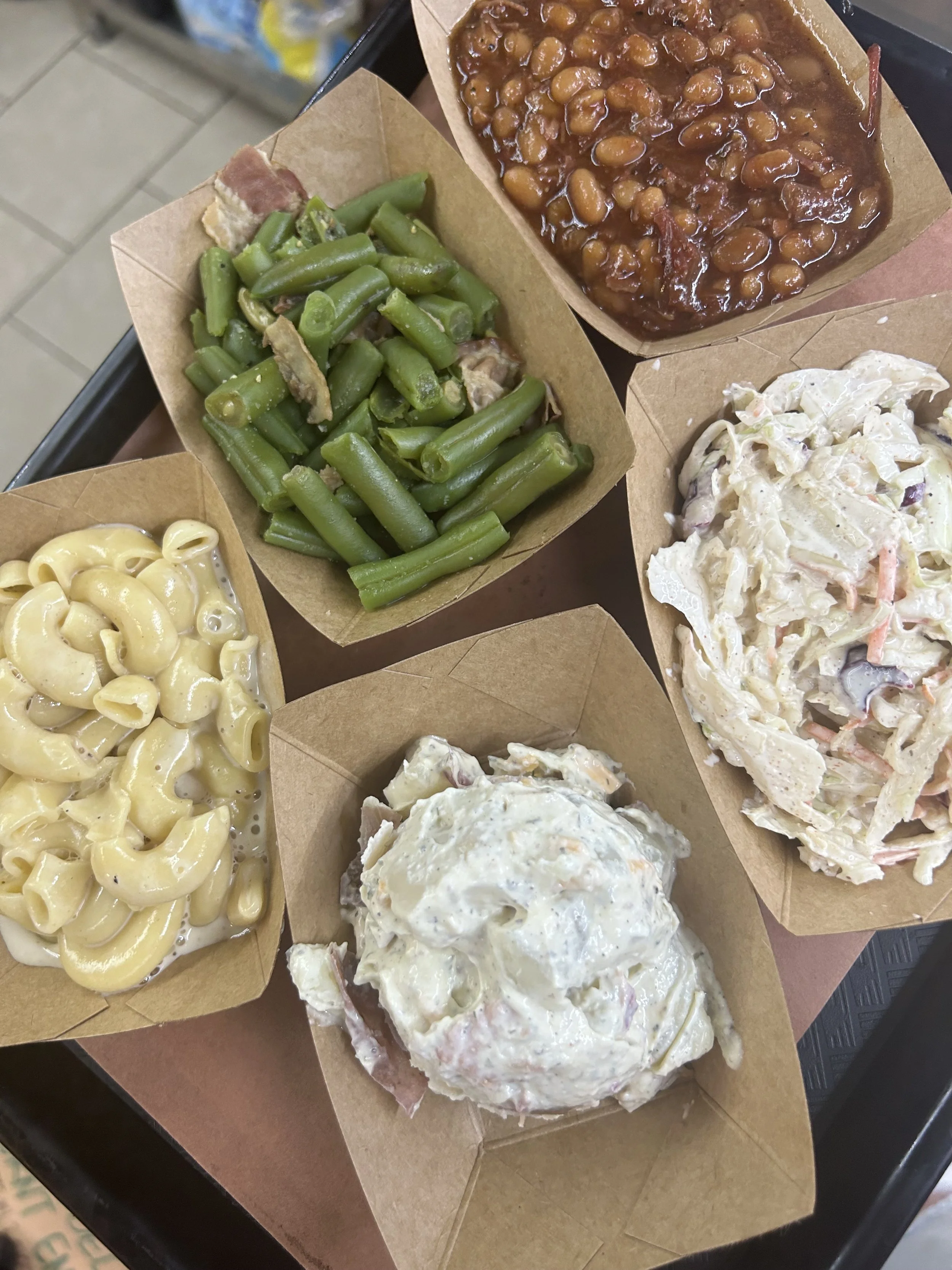 Fast food meal with macaroni and cheese, green beans, baked beans, and coleslaw served in small paper trays.