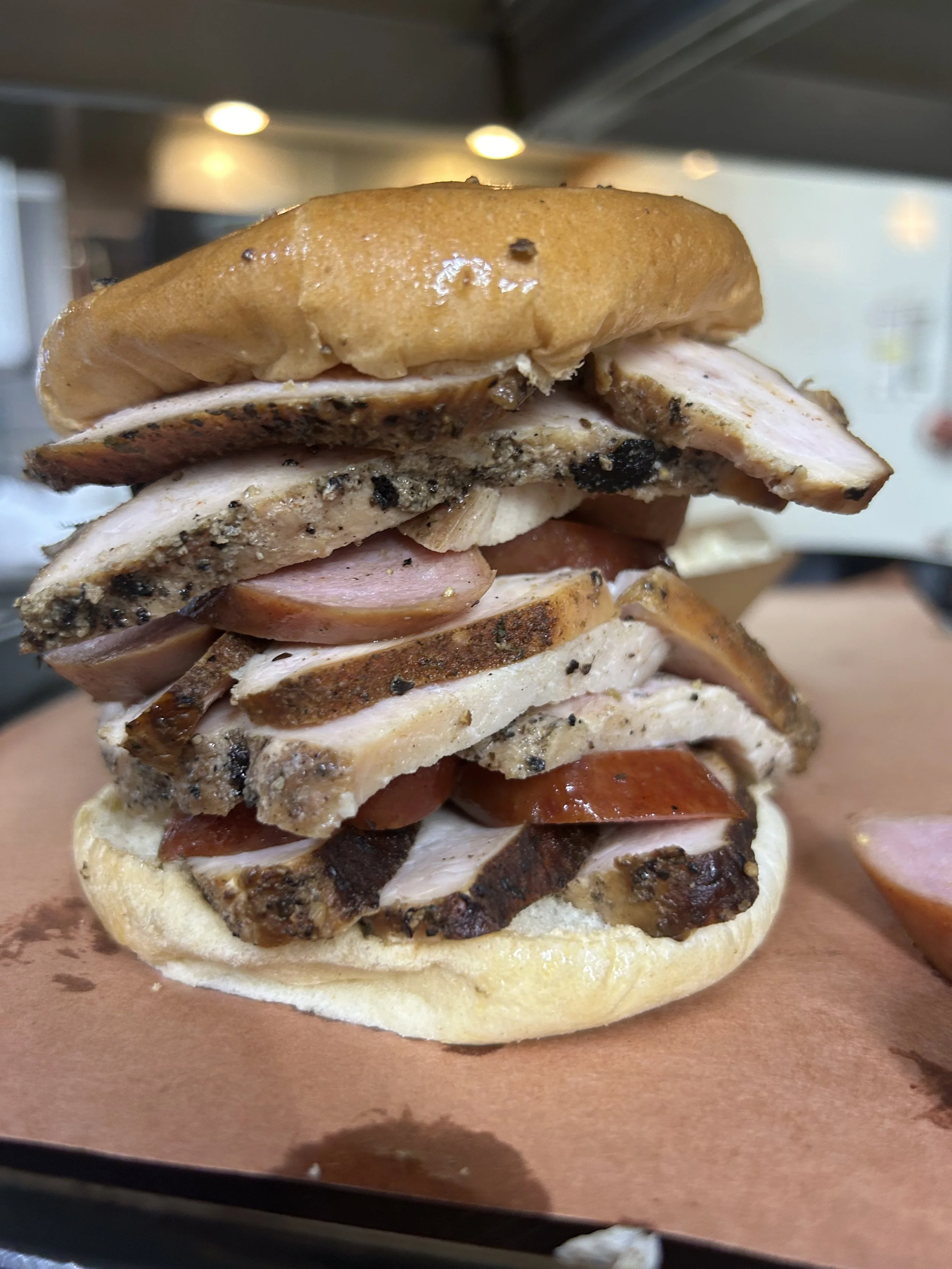 Close-up of a hamburger bun topped with sliced turkey, ham, and peppered chicken or turkey, with some seasoning visible, on a paper-lined tray.