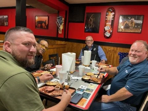 Four men sitting around a table in a restaurant, enjoying a meal with sandwiches, fries, and drinks.