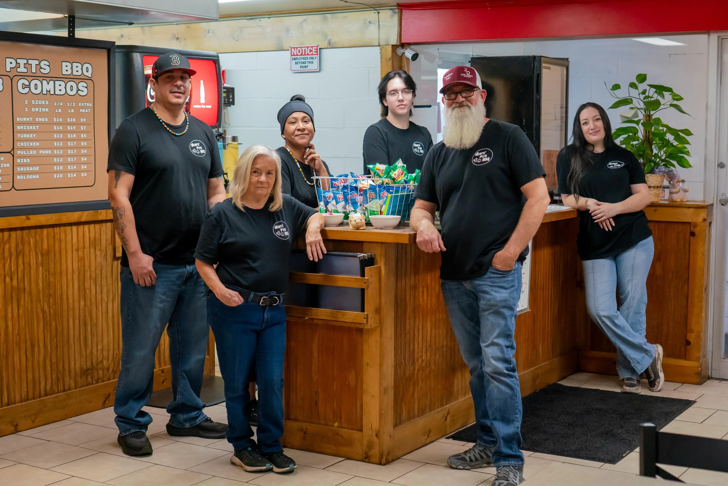Group of six people standing inside a restaurant near a counter with a menu board on the wall. Four of them are wearing black shirts with a logo, and one woman is wearing a dark beanie. Behind the counter, there are snack bags and bowls.