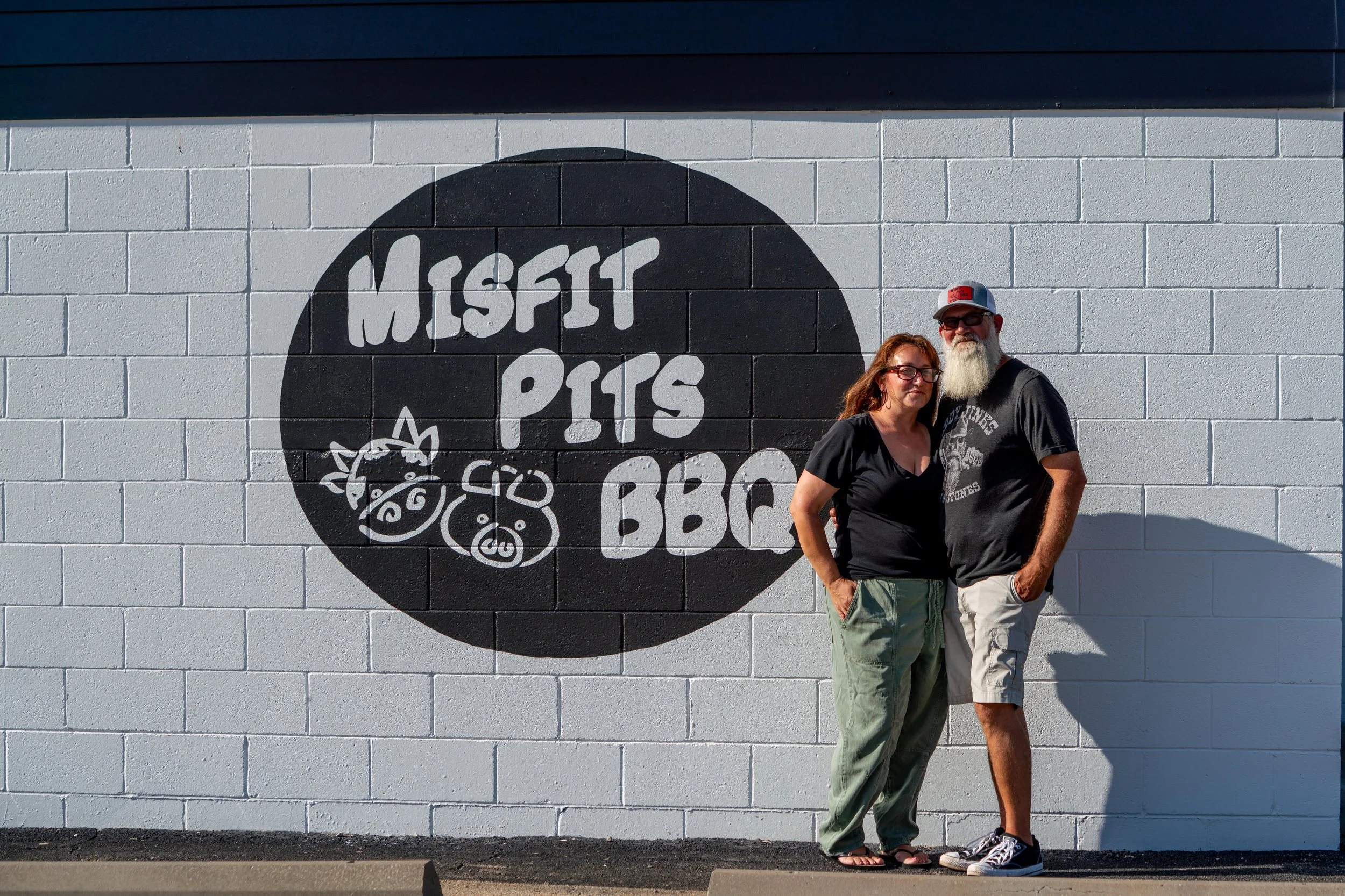 A man and woman standing together in front of a large black and white mural on a brick wall that reads 'MISTI PITS BBQ'. The man has a long white beard, sunglasses, a gray cap, and is wearing a black T-shirt and light-colored shorts. The woman has red hair, glasses, and is wearing a black shirt and green pants.