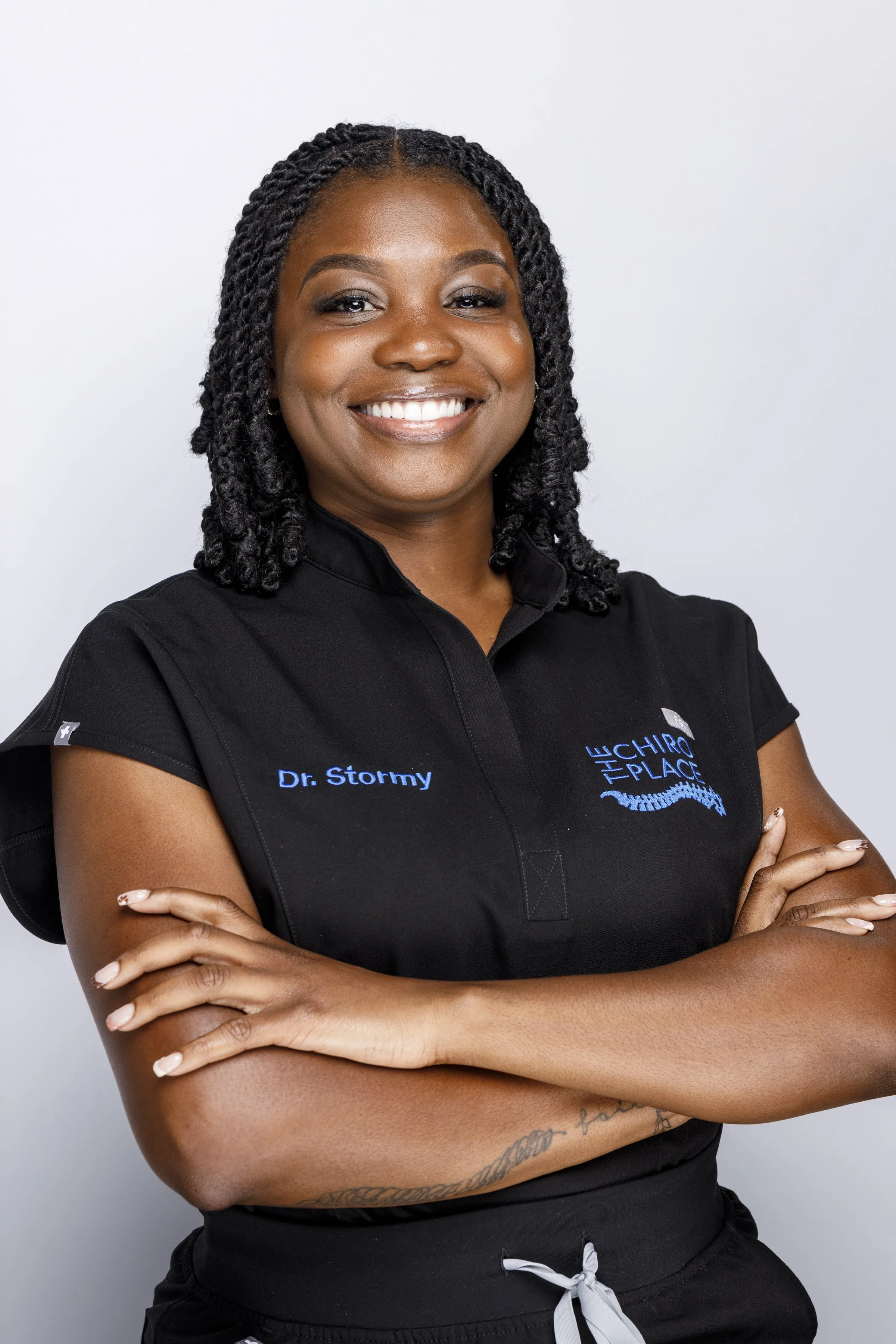 A smiling woman with dark hair styled in twists, arms crossed, wearing a black shirt with embroidered name and logo, standing against a white background.