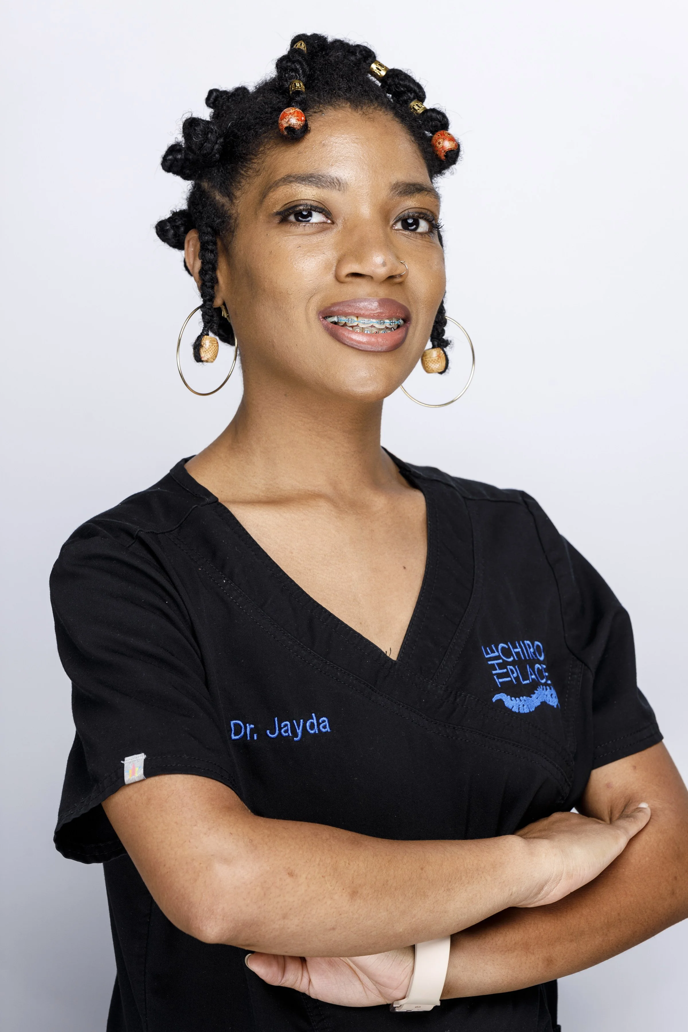 Portrait of a woman with styled hair adorned with decorative beads, wearing braces, large hoop earrings, and a black scrubs top with her arms crossed, standing against a plain light gray background.