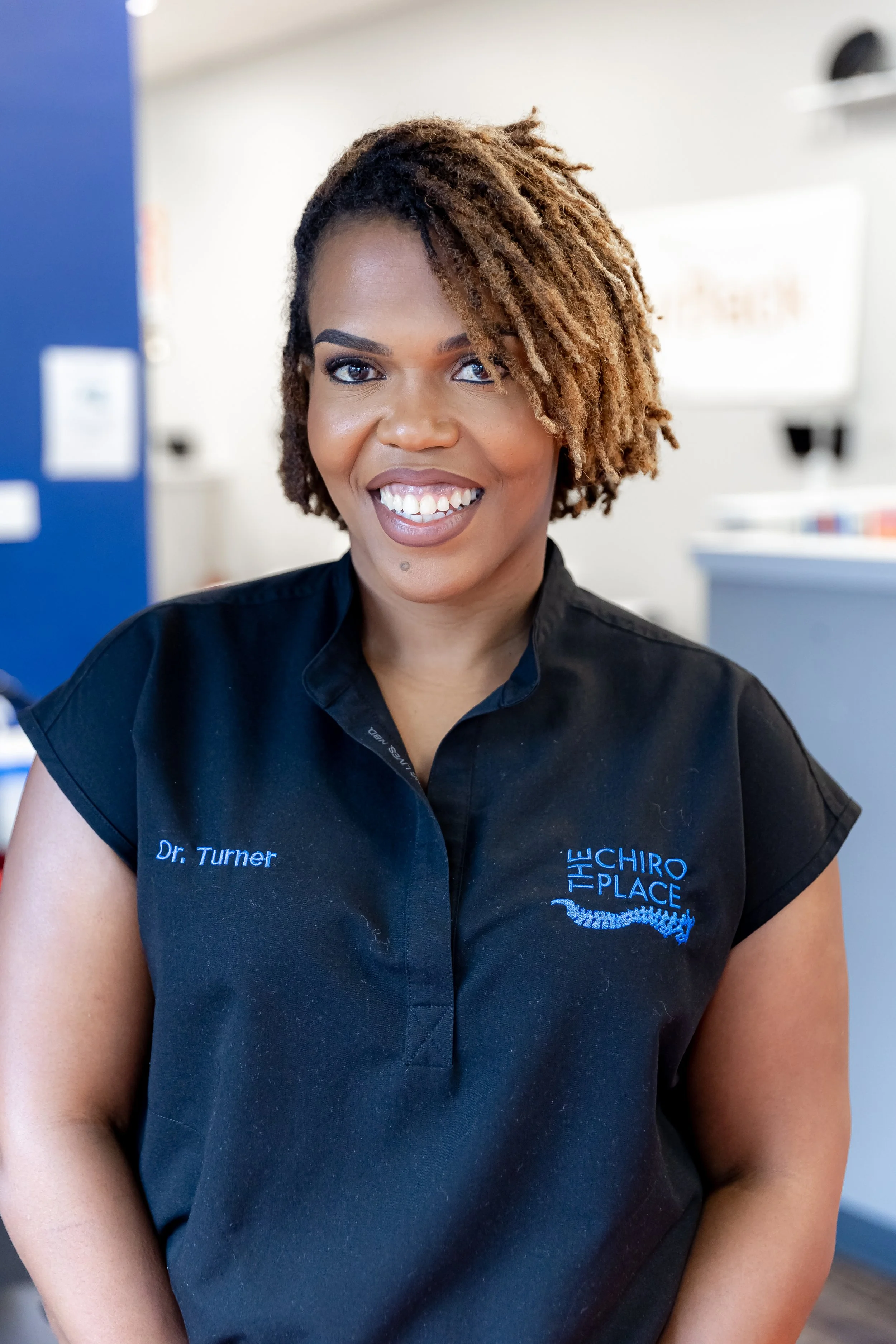 A woman with short, curly, two-tone hair and dark eyes smiling, wearing a black uniform with 'Dr. Turner' and 'The Chiro Place' embroidered on it, inside a clinic or dental office.