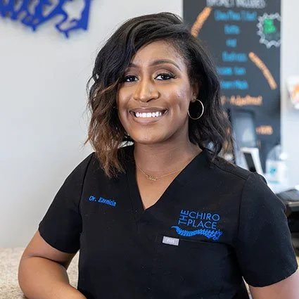 A woman with dark, wavy hair and hoop earrings smiling in a black medical uniform with "The Chiro Place" logo, standing in a professional setting.