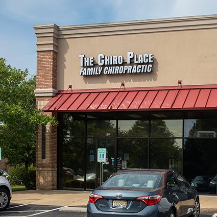 Front view of The Chiro Place Family Chiropractic office with a parking lot and cars, including a gray car in the foreground, and a tree on the left side.