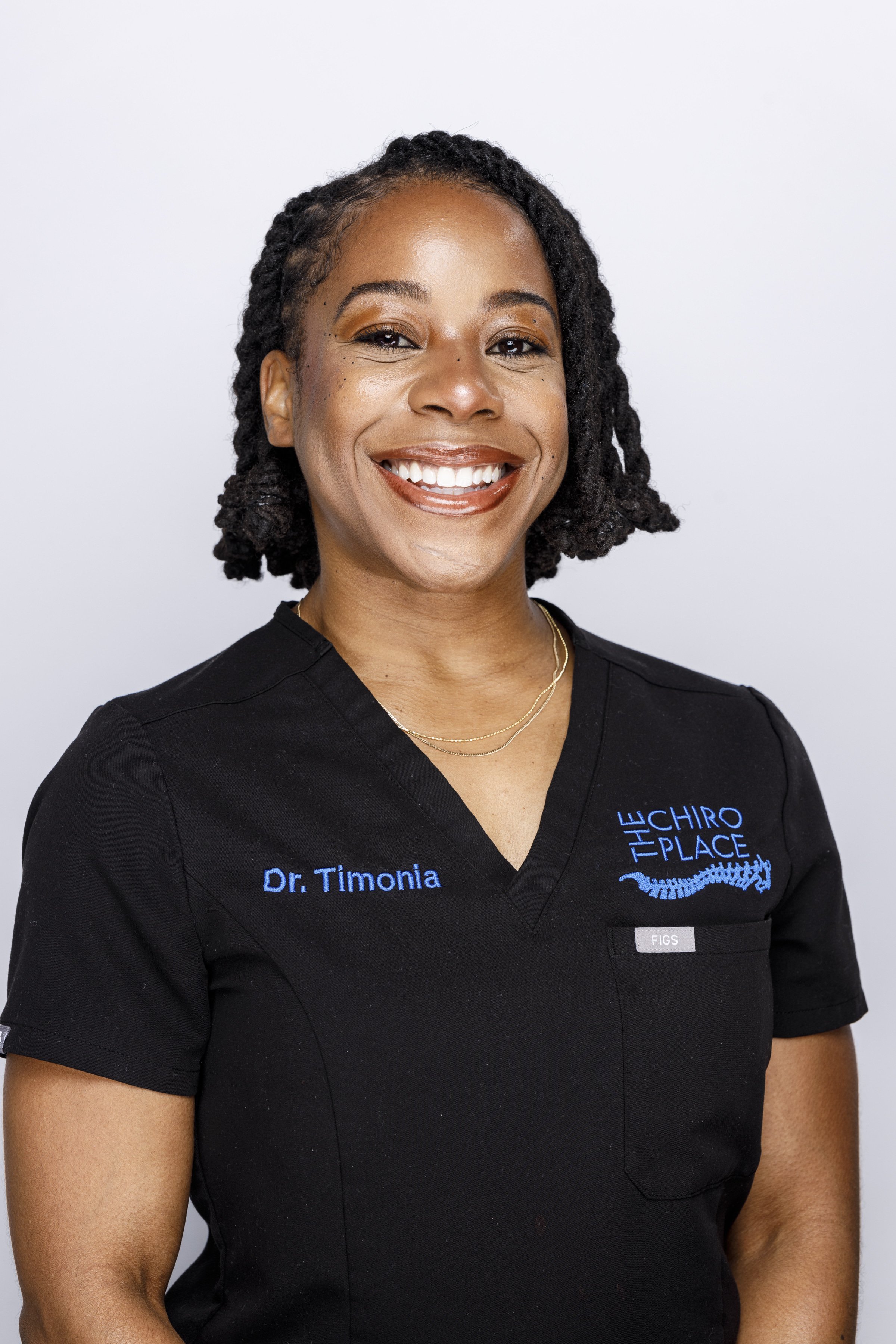 A woman with short curly hair wearing a black medical scrub top with blue embroidered text that says 'Dr. Timonia'. She is smiling and standing against a plain light gray background.
