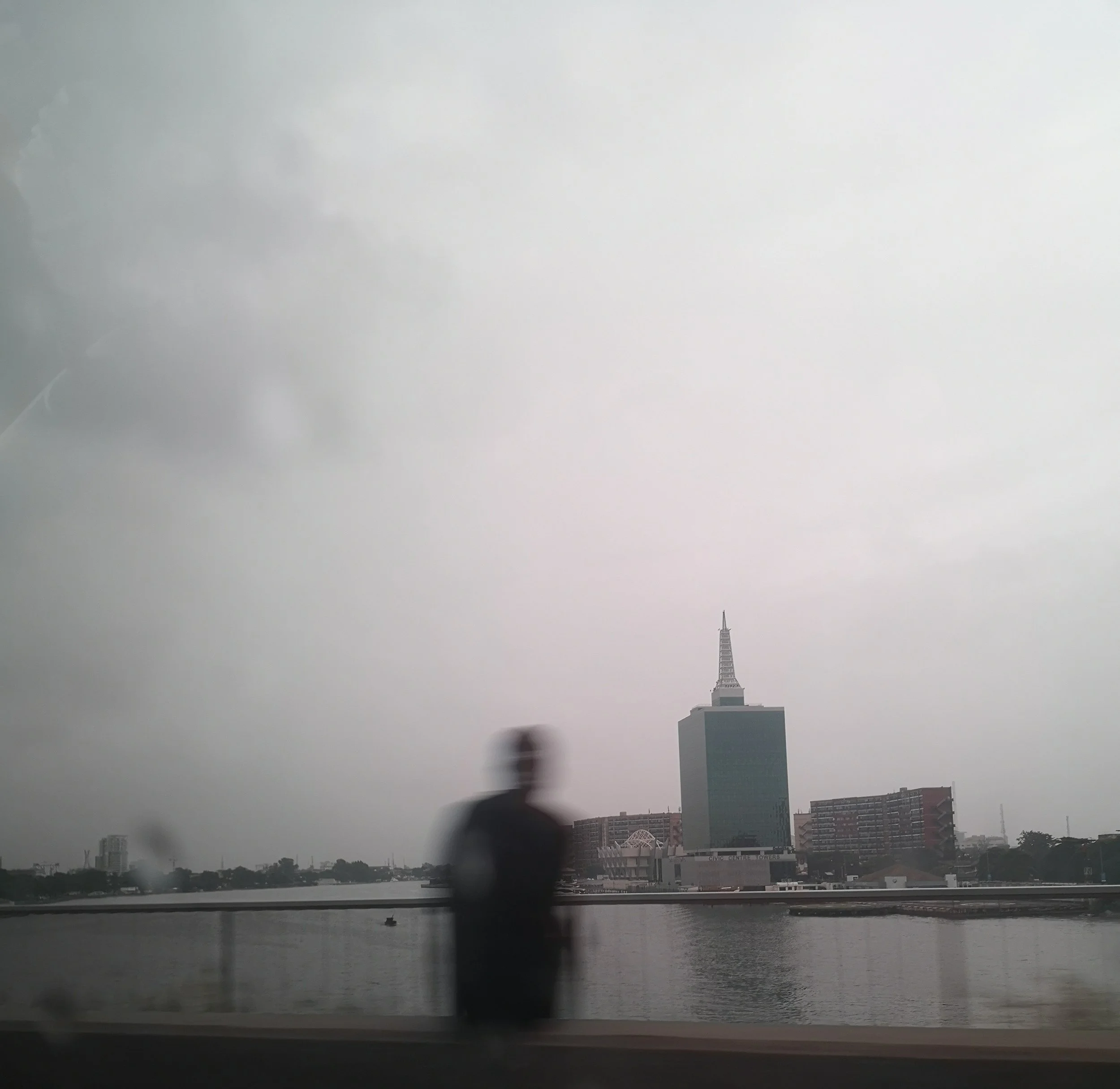 Cityscape with a large body of water in the foreground. A bridge is seen, and a skyline with numerous buildings including a tall, pointed tower in the background on a cloudy day.