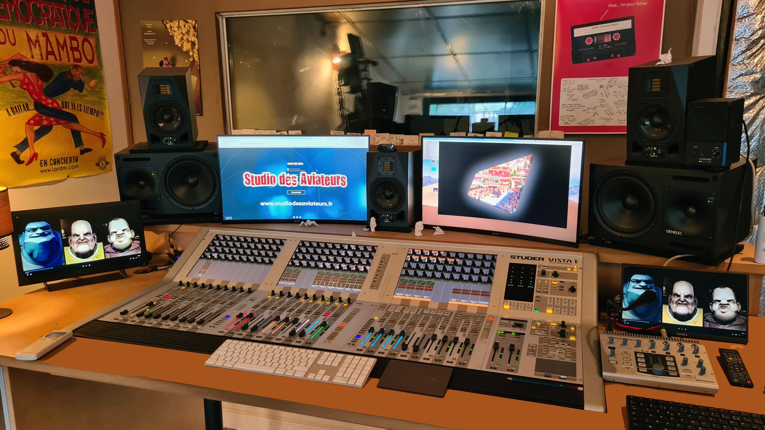 Studio d'enregistrement avec deux écrans d'ordinateur, un console de mixage, des haut-parleurs et divers équipements audio. Sur un écran, il y a un logo de 'Studio des Aviateurs' et un site web. La pièce est décorée avec des posters colorés, dont un 