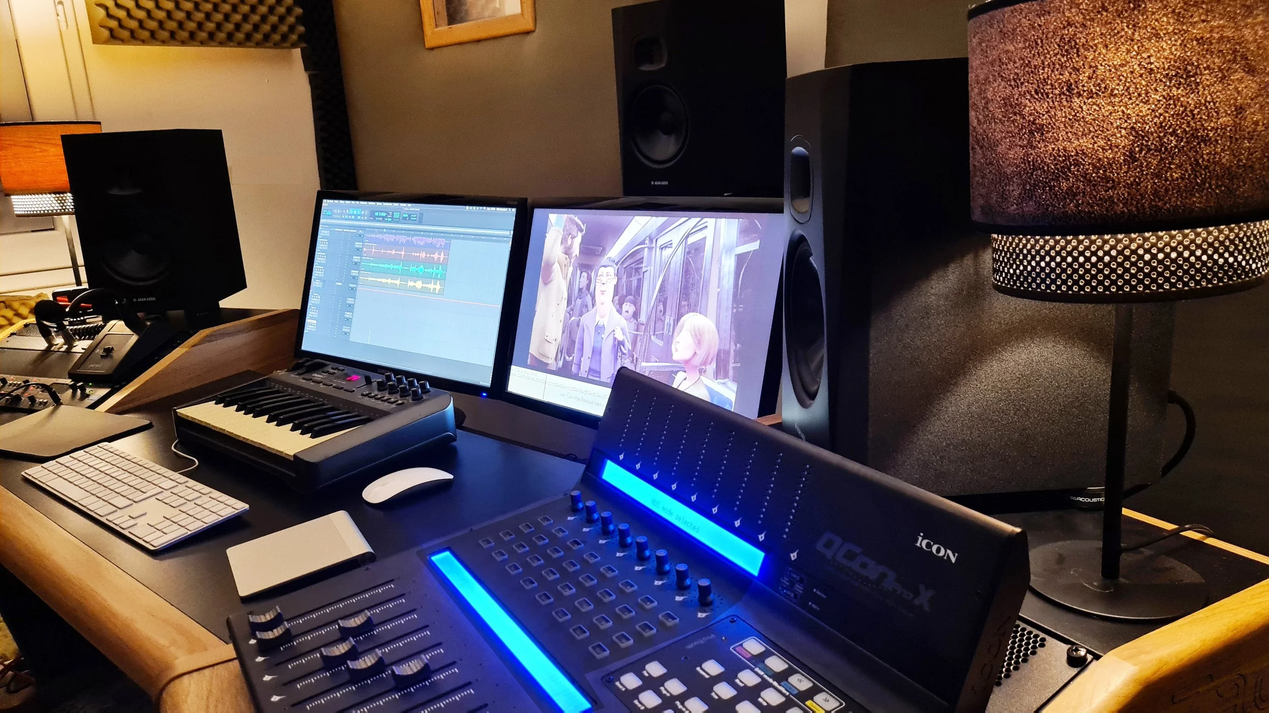 Un studio d'enregistrement avec deux écrans, un clavier, une console de mixage avec diodes bleues, deux haut-parleurs et une lampe sur pied. Sur un écran, on voit un logiciel de musique et sur l'autre, un film d'animation ou une vidéo. Le studio disp