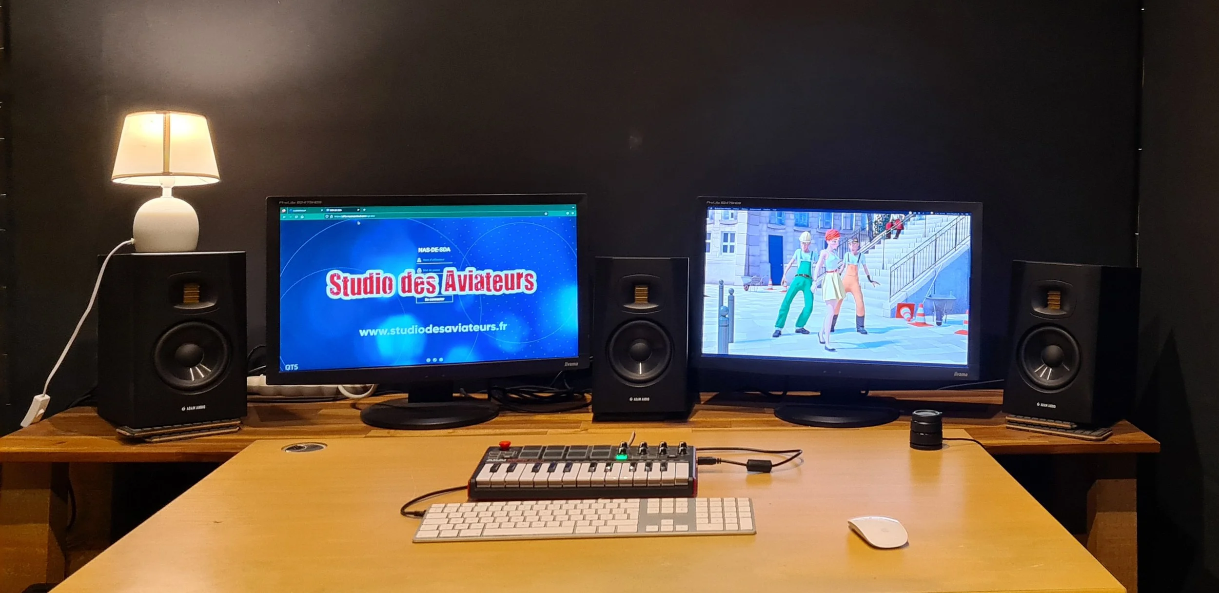 Un espace de bureau avec deux écrans d'ordinateur, des haut-parleurs, une lampe, un clavier, une souris et une petite machine de musique, dans une pièce sombre.