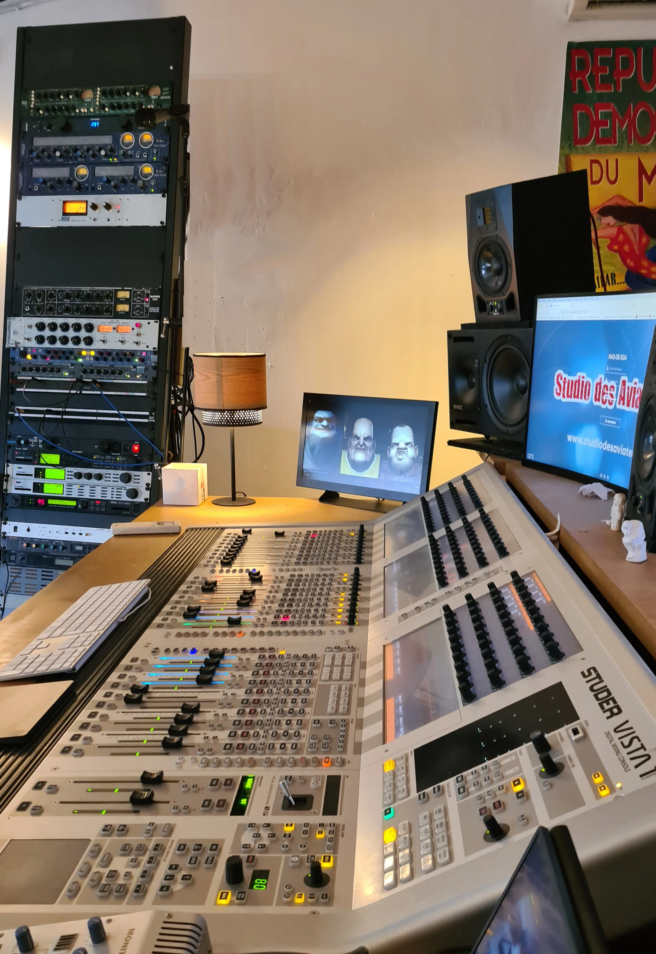 Une salle d'enregistrement audio avec une console de mixage, des haut-parleurs, deux écrans d'ordinateur, un lampadaire, et une affiche à l'arrière-plan