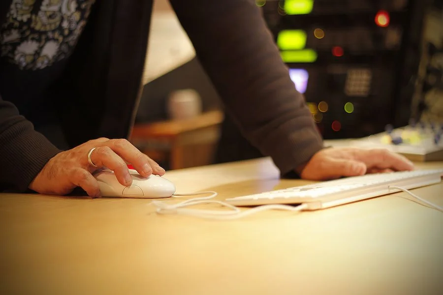 Main d'une personne utilisant une souris d'ordinateur et une autre main reposant sur un clavier sur une table, avec un fond flou d'équipements électroniques.