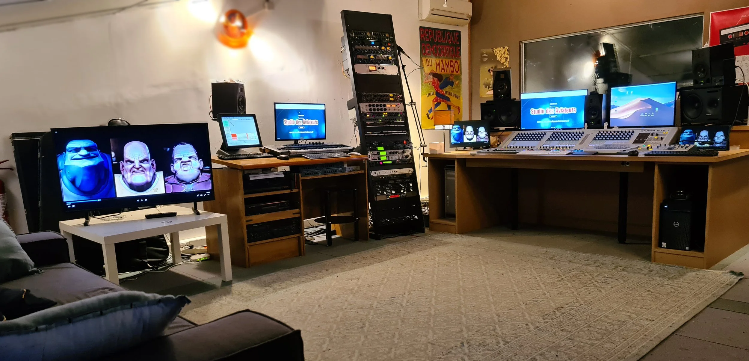 Studio d'enregistrement audio avec plusieurs écrans, consoles de mixage, haut-parleurs, et équipement électronique, décoré avec un poster coloré sur le mur.