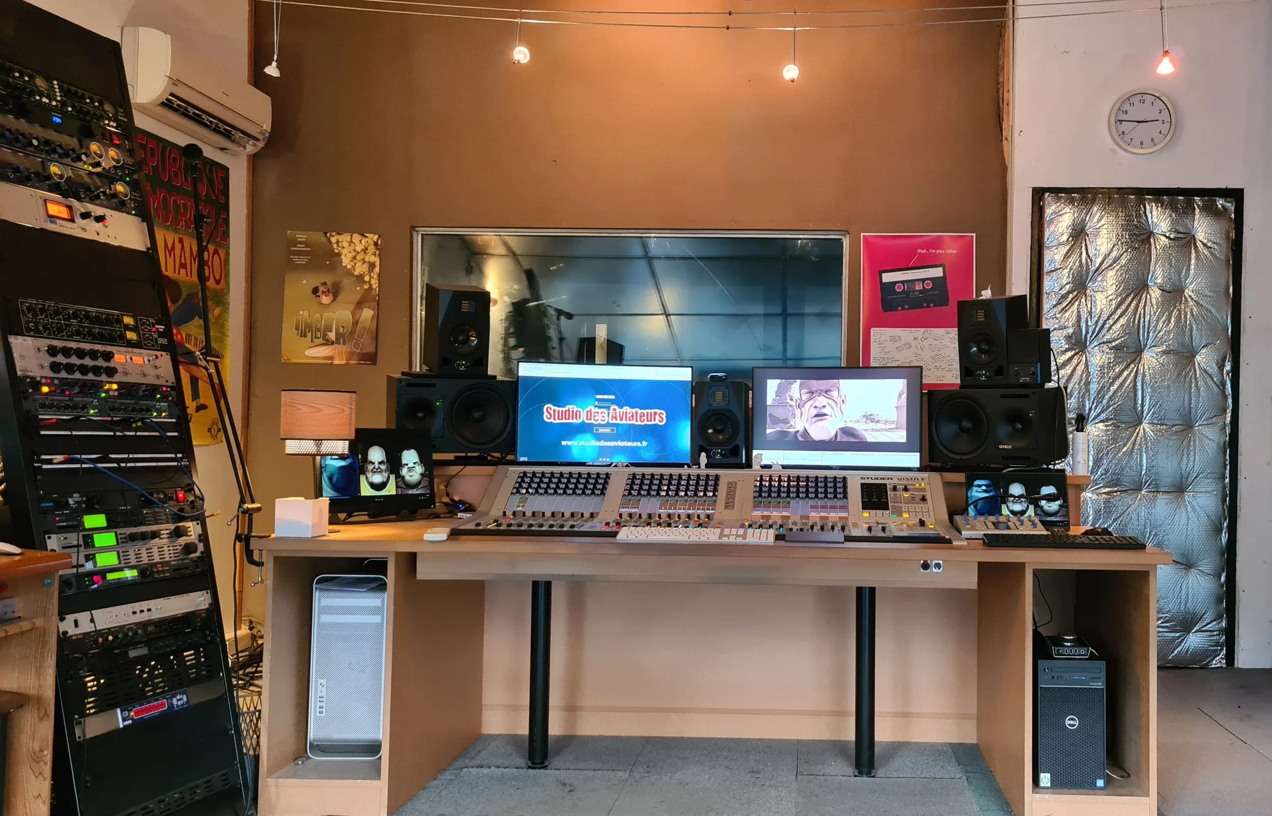 Studio d'enregistrement avec des équipements audio professionnels, écrans d'ordinateur, mobilier en bois, posters sur le mur, et un mur recouvert d'isolant acoustique.