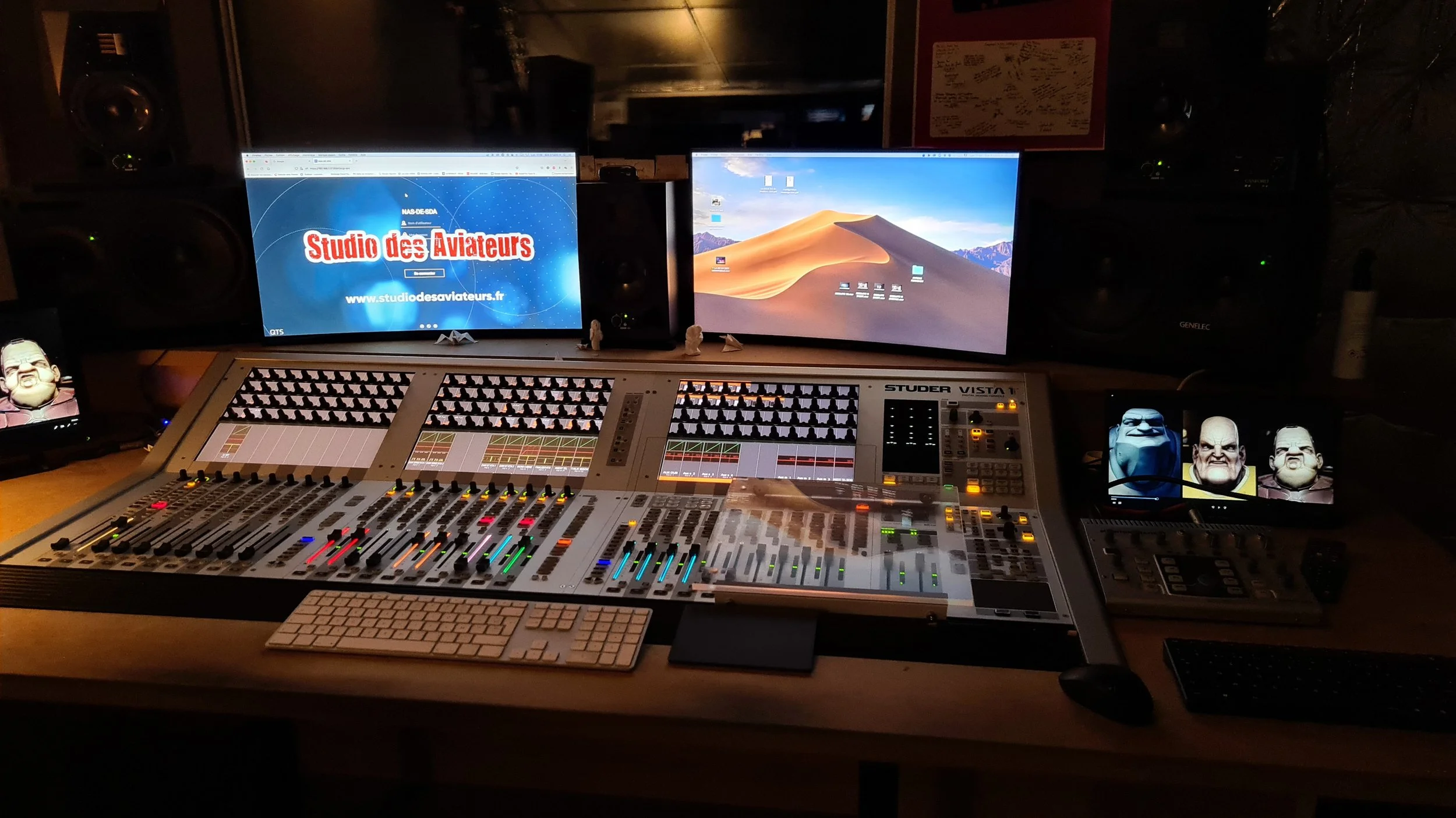 Photo d'un poste de travail en studio d'enregistrement avec deux écrans, une console de mixage, des haut-parleurs, plusieurs figurines et un clavier, dans une pièce sombre.