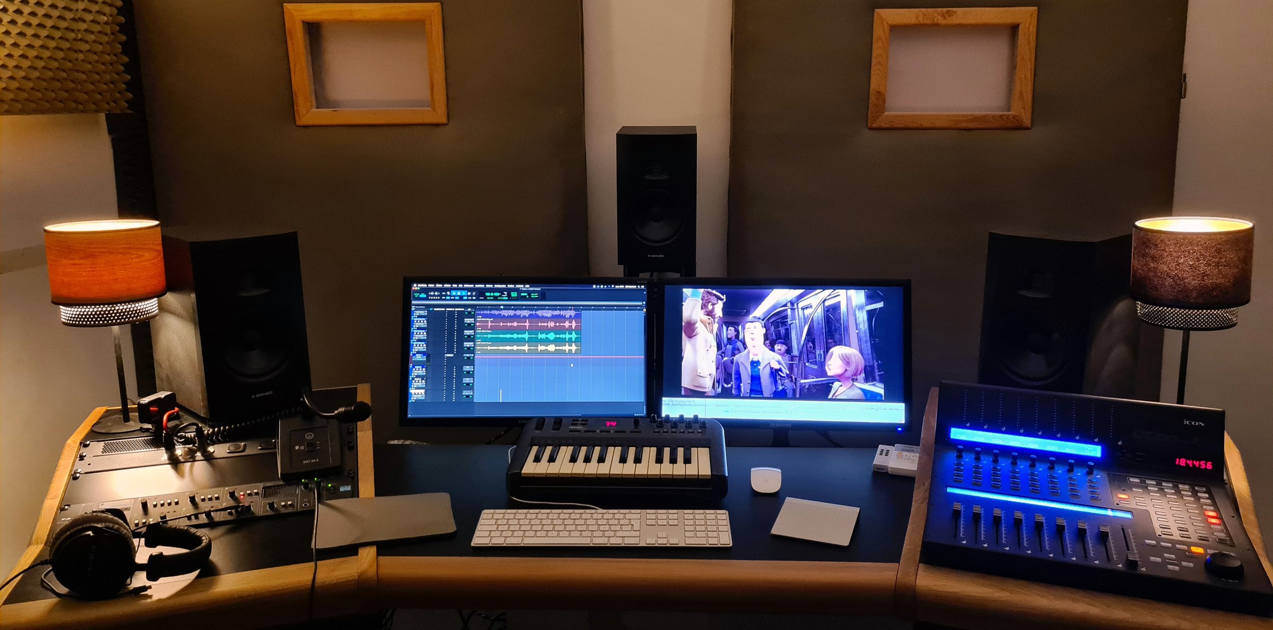 Studio d'enregistrement avec deux écrans affichant un logiciel de montage vidéo et une scène de film, deux haut-parleurs, une console de mixage, un clavier MIDI, une souris, un casque audio, des lampes de lecture, et des murs insonorisés.