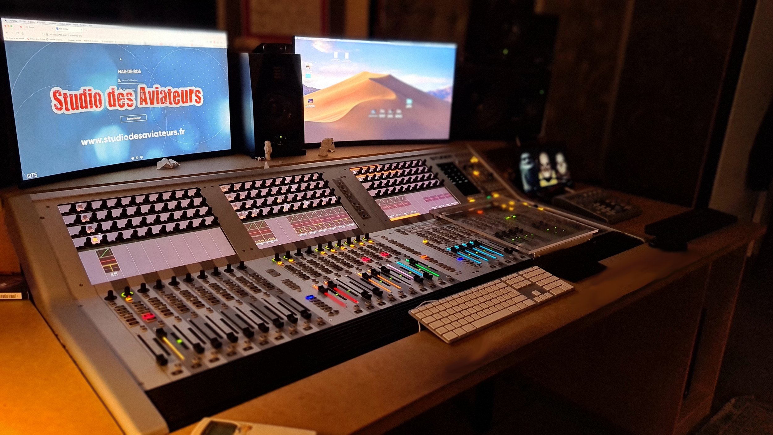 Studio d'enregistrement audio avec console de mixage, deux écrans d'ordinateur, un smartphone, et divers équipements de production sonore.