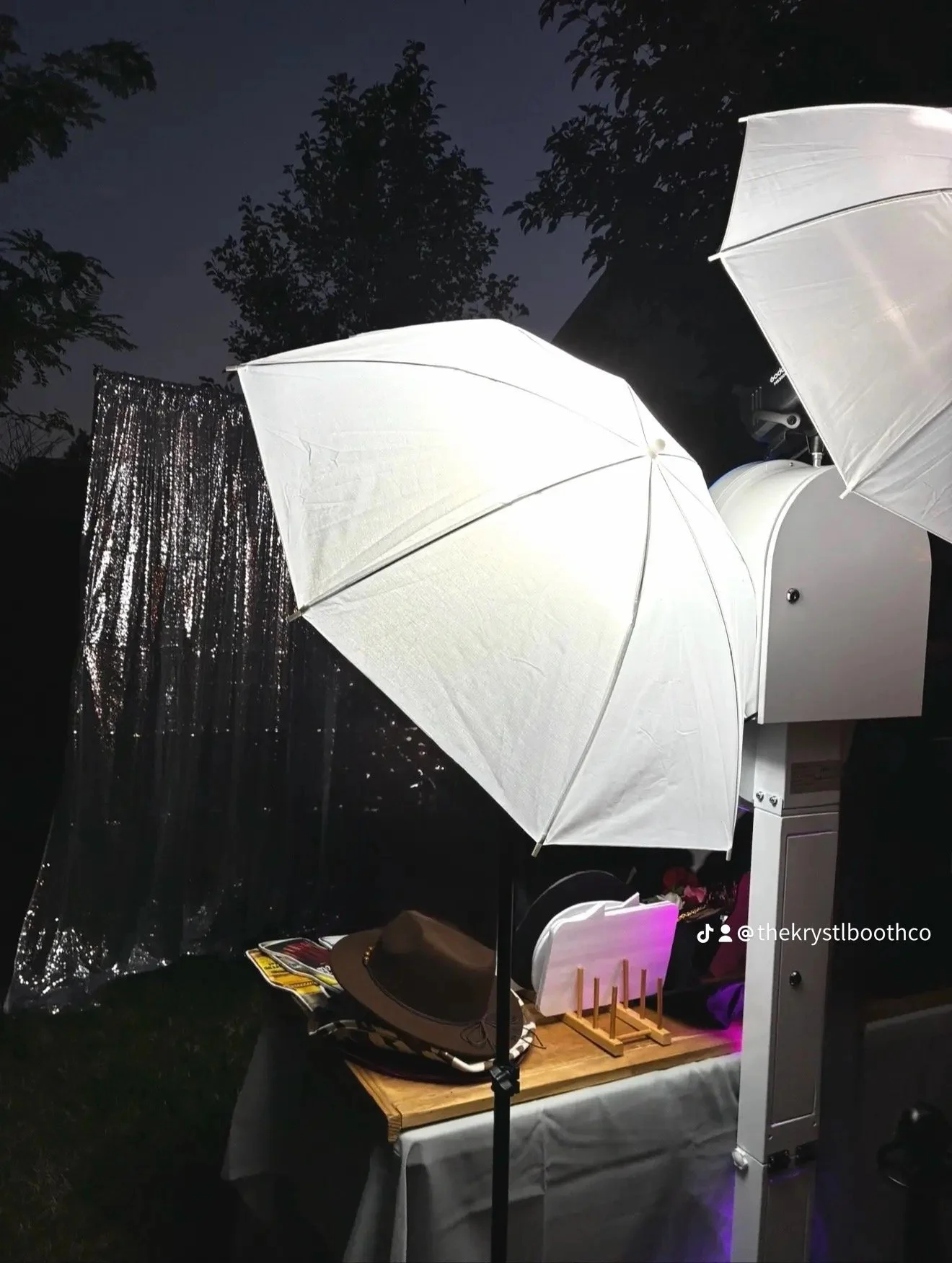 A photo booth setup with two large white umbrellas, a table with various props including hats, and a backdrop with shiny black streamers, outdoors during nighttime.