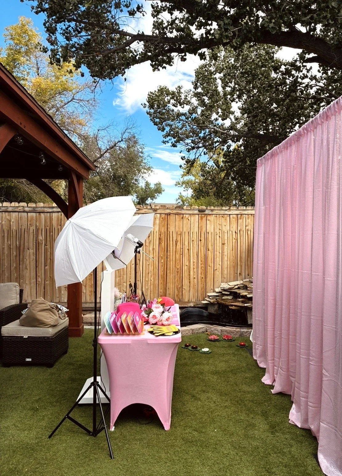 Outdoor backyard setup with photography equipment, including a white umbrella and lighting, a pink table with paper plates, flowers, and utensils, a pink curtain backdrop, and a wooden fence in the background. The scene is on a grassy area with trees