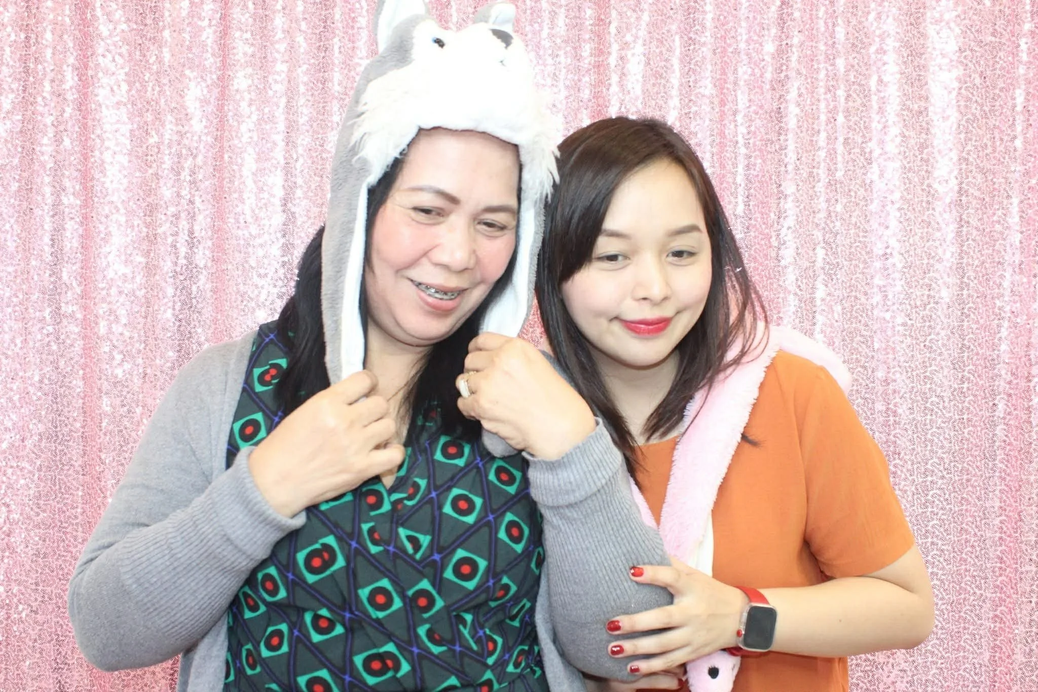 Two women standing close together in front of a pink sequined backdrop. The woman on the left is wearing a Hello Kitty hat and a patterned top, while the woman on the right is wearing an orange shirt with pink trim and a pink hooded garment, smiling 