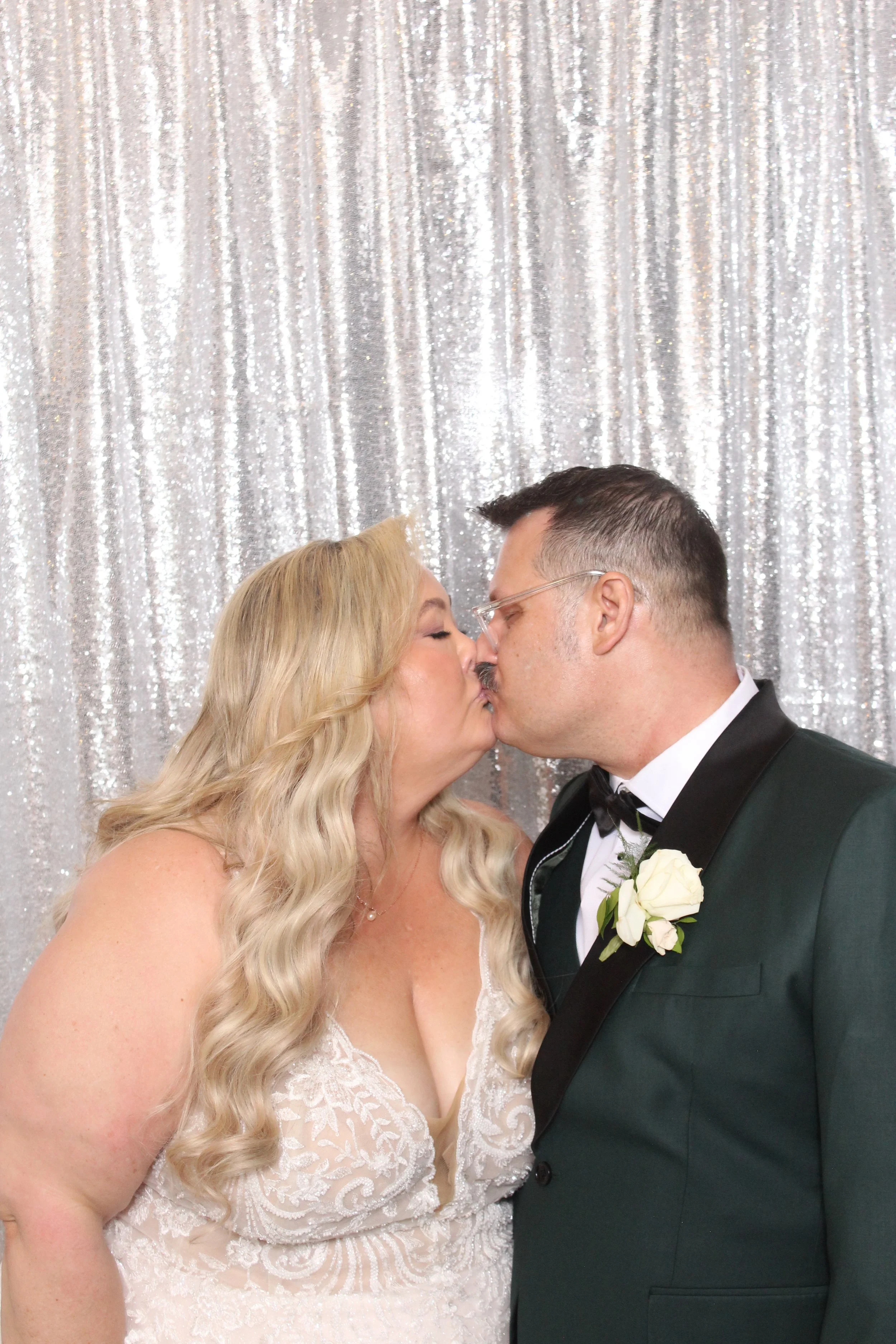 A bride and groom sharing a kiss at their wedding, with a sparkly silver curtain backdrop.