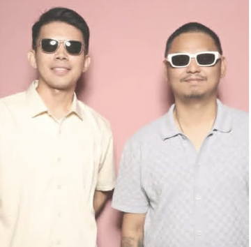 Two men wearing sunglasses and light-colored shirts standing against a pink background.