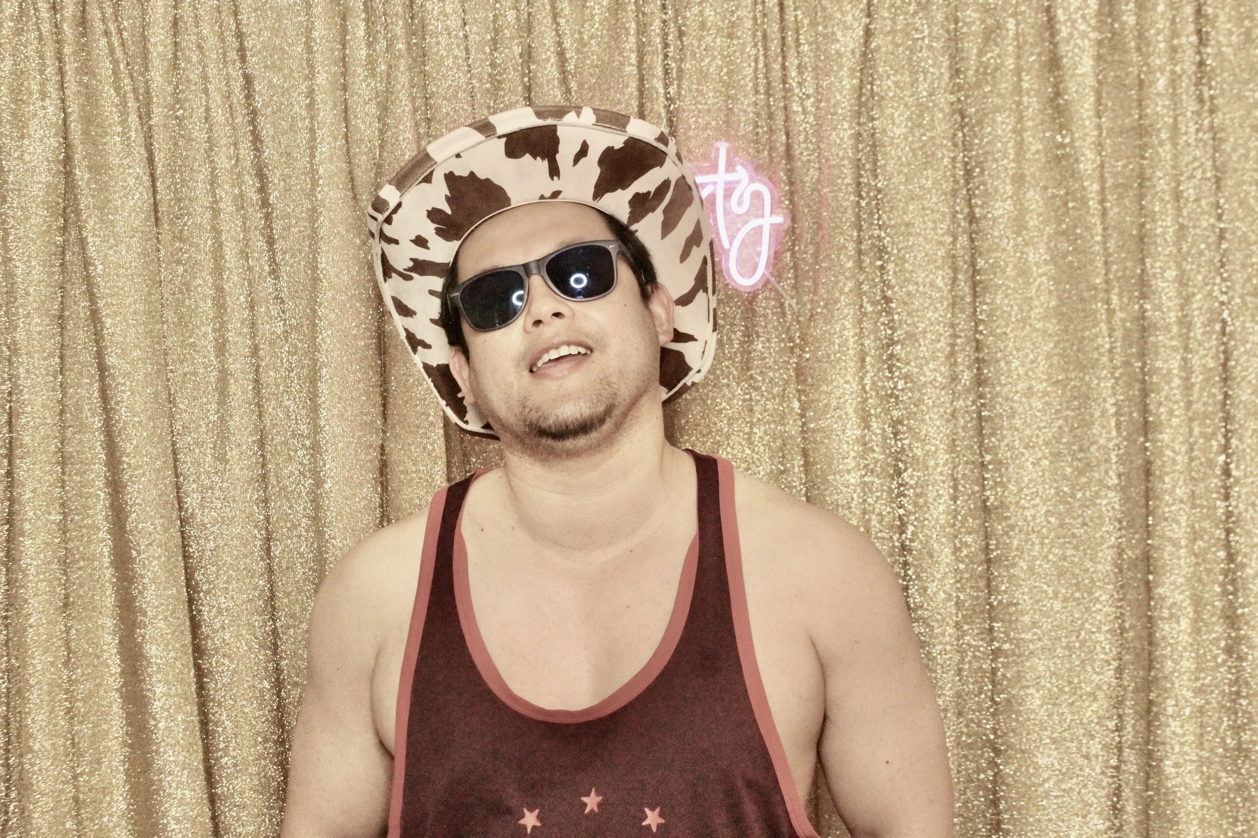 Person wearing a cowboy hat and sunglasses, in front of a gold glittery background with a pink neon sign.