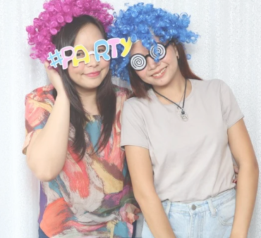 Two women wearing colorful wigs and holding a sign that says "#PARTY" at a celebration or party.