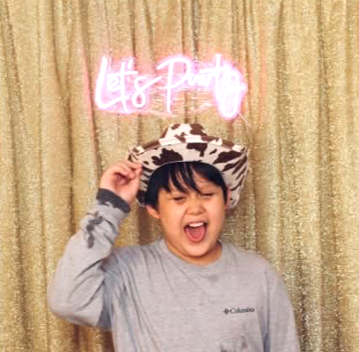 A young boy wearing a cowboy hat, smiling with his eyes closed, standing in front of a gold curtain with a pink neon sign that says 'Let's Party'.
