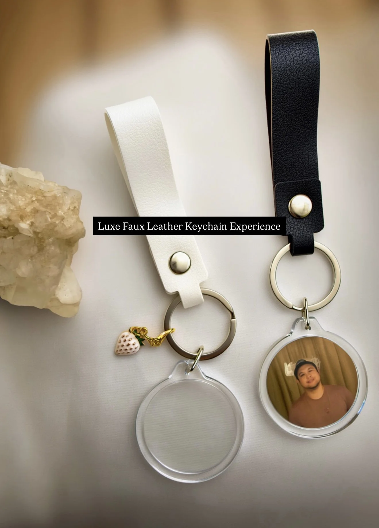 Two faux leather keychains, one white with a silver button and one black with a silver button, laying next to a rock, with the text 'Luxe Faux Leather Keychain Experience' captioned across. The keychains have round photo and transparent tags attached.