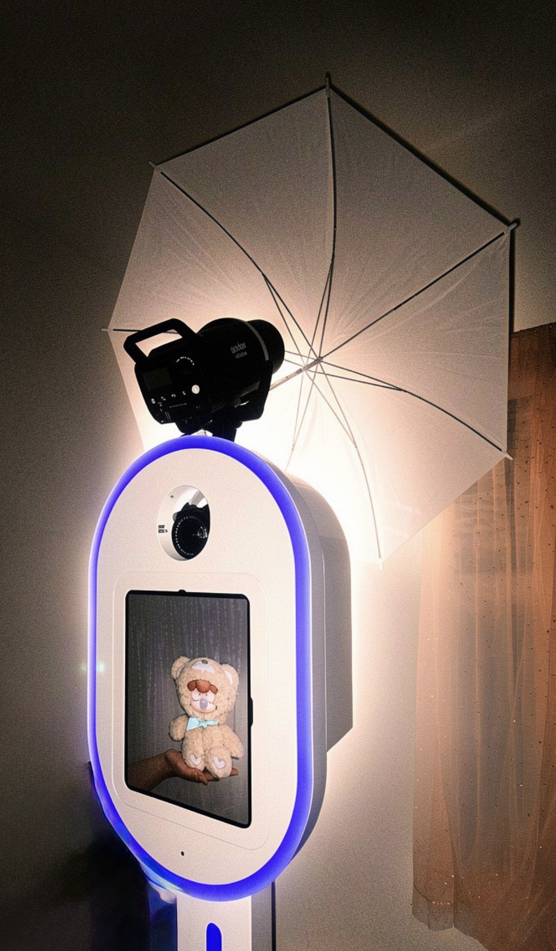 A teddy bear plush toy holding a smaller teddy bear plush toy in front of a mirror with a person taking a photo. There is an umbrella light attached to a device with a circular screen and adjustable head, all on a white table with a blue LED streak.