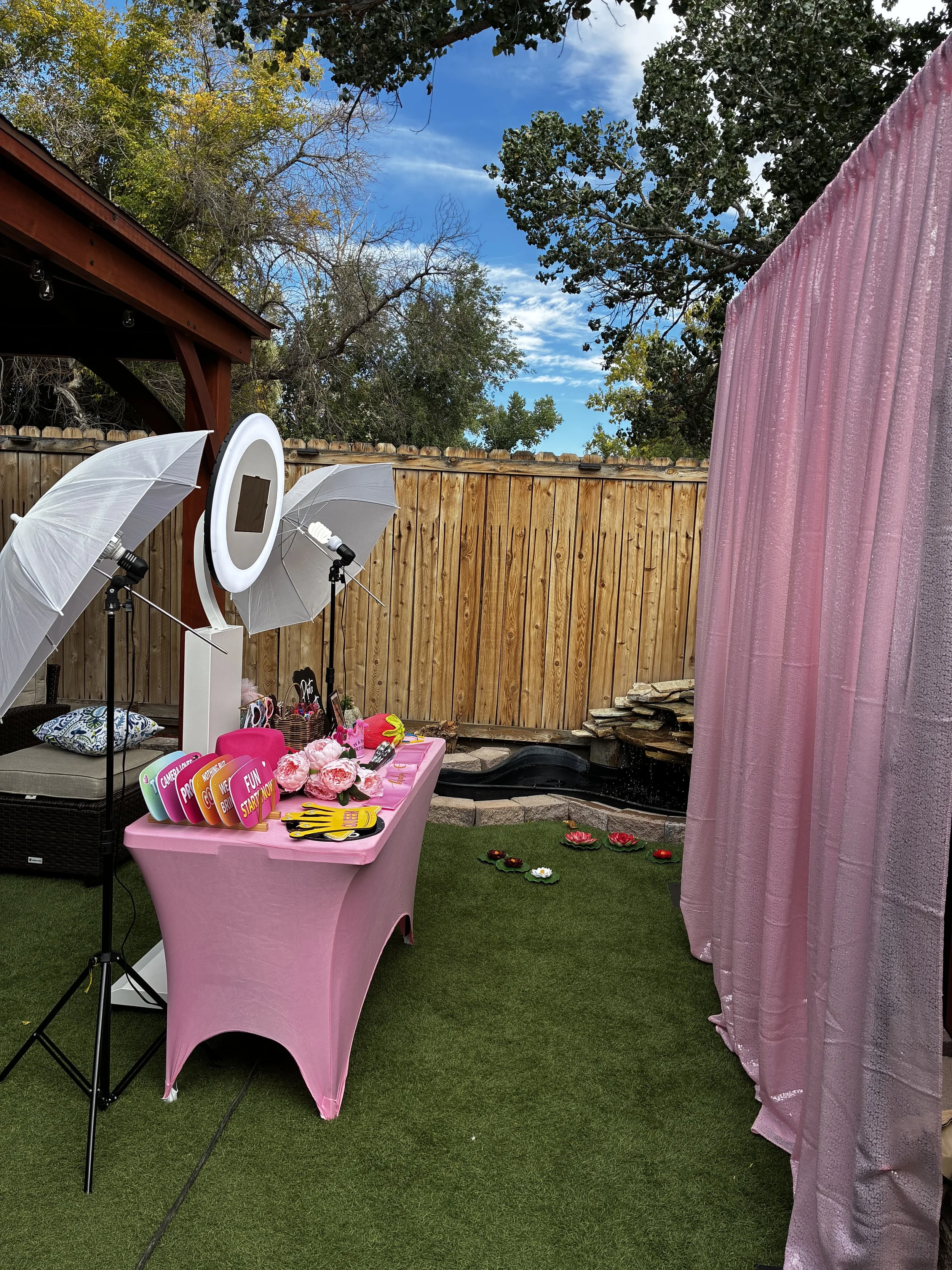 Decorative outdoor party setup with a pink table, colorful foam photo props, a pink curtain, and photo booth accessories, with a wooden fence, trees, and a cloudy sky in the background.