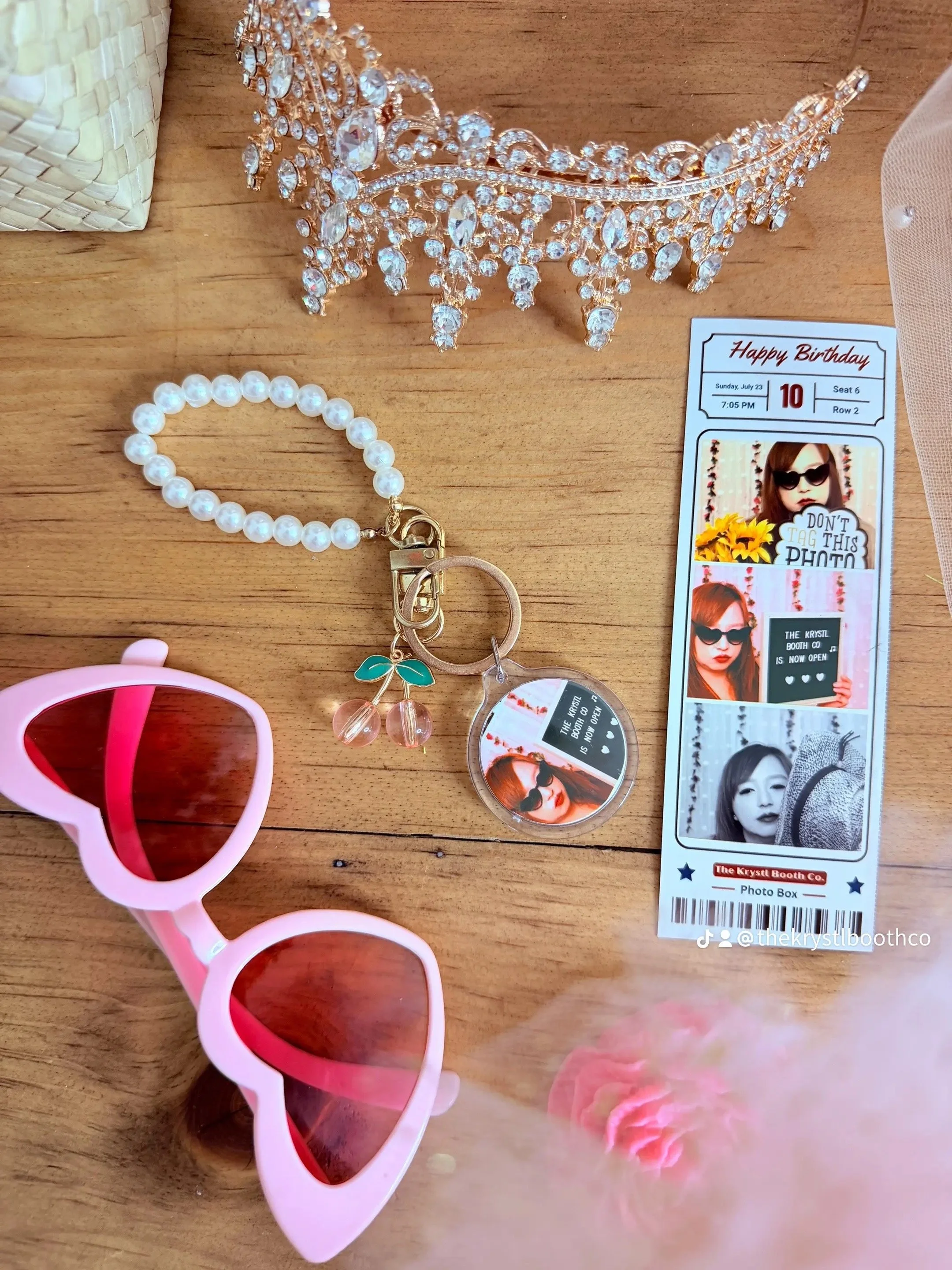 Pink heart-shaped sunglasses, a pearl bracelet with a cherry charm, a rhinestone tiara, a photo booth strip with black-and-white and color photos, and a birthday ticket card on a wooden surface.