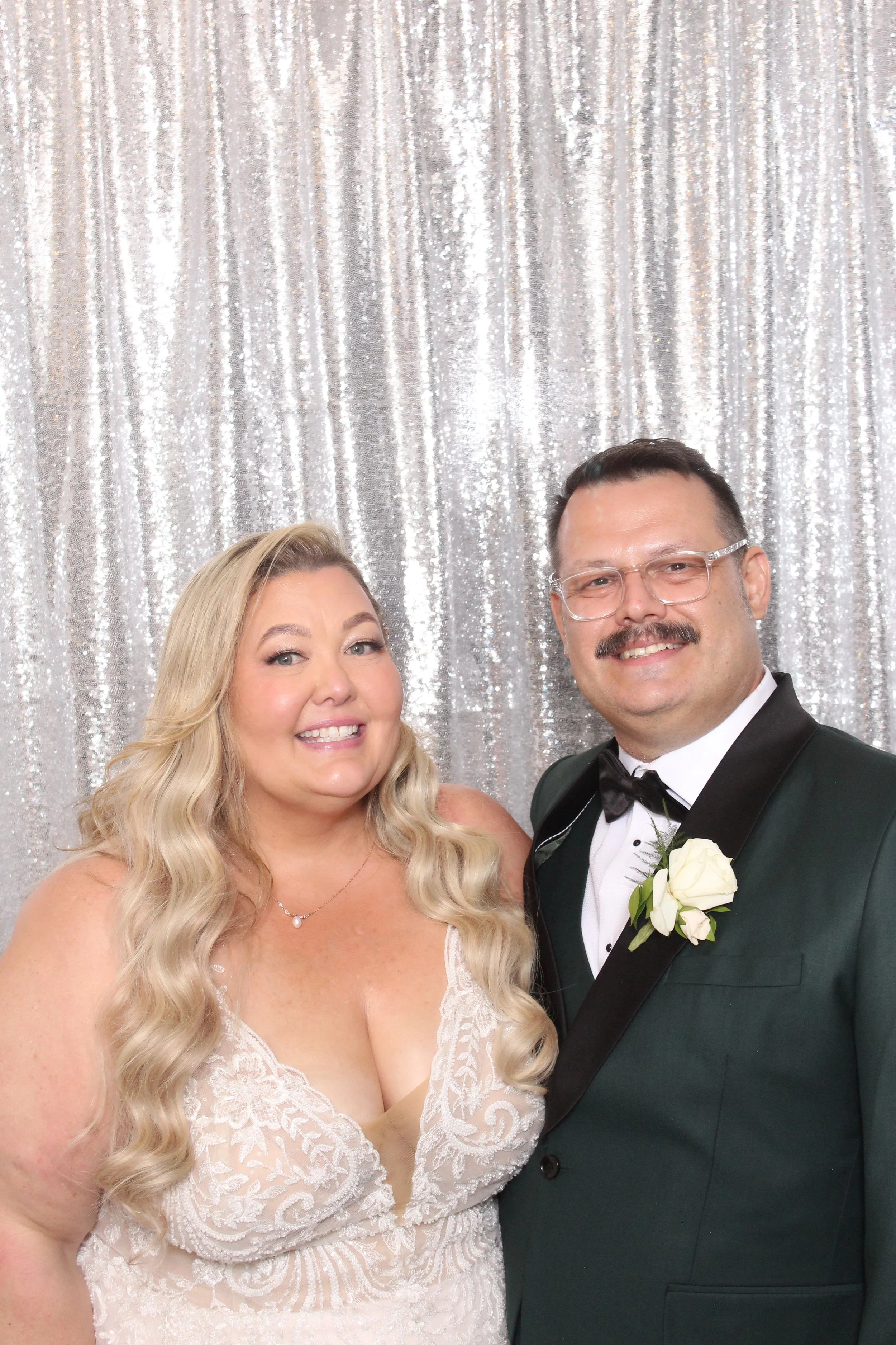 A couple dressed in wedding attire standing in front of a sparkly silver backdrop. The woman has long, wavy blonde hair and is wearing a white lace wedding dress. The man is wearing a black tuxedo with a white shirt, black bow tie, and glasses. He ha