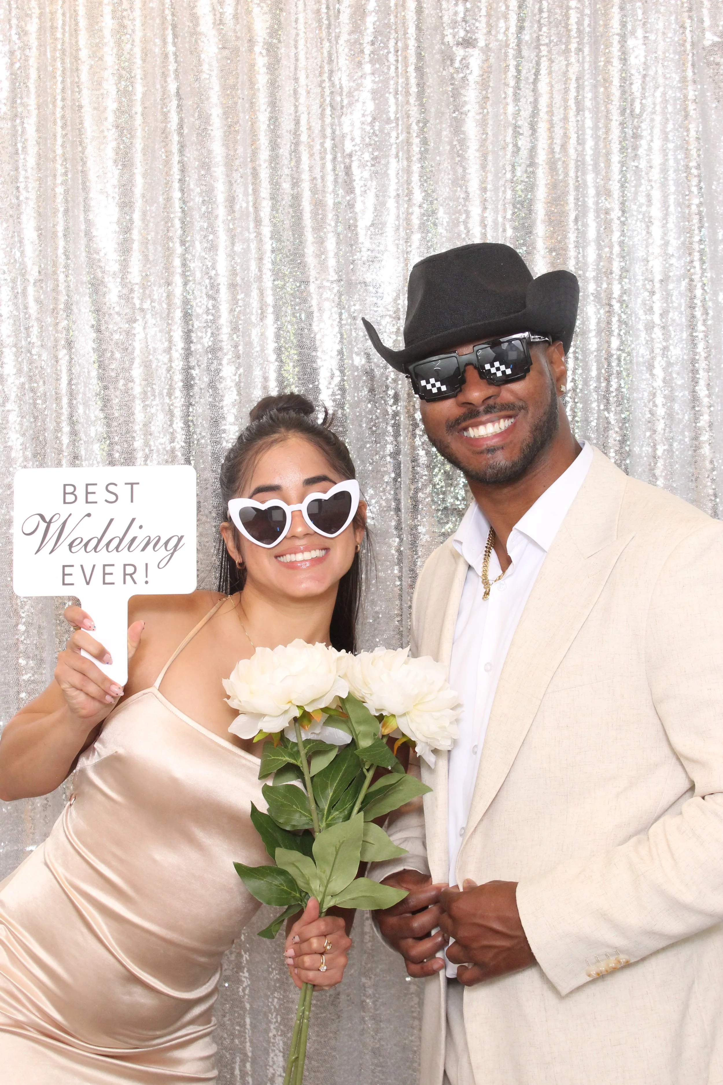 A smiling woman in a satin dress holding a bouquet of white flowers and a sign that reads "Best Wedding Ever!" alongside a smiling man in a beige suit and black cowboy hat, both wearing funny pixelated sunglasses, standing in front of a silver sequin