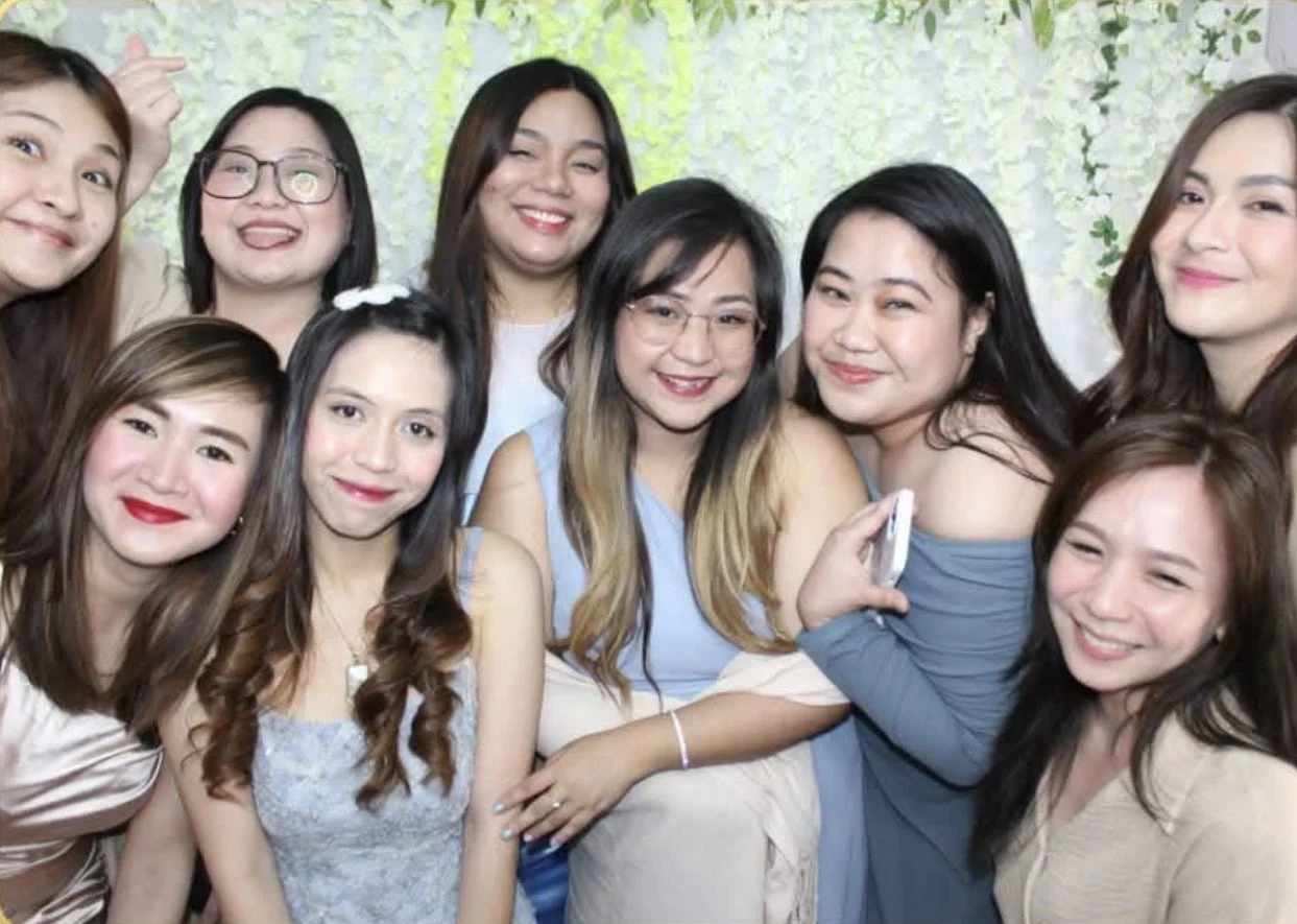 Group of nine women smiling at a social gathering, with a green leafy background.